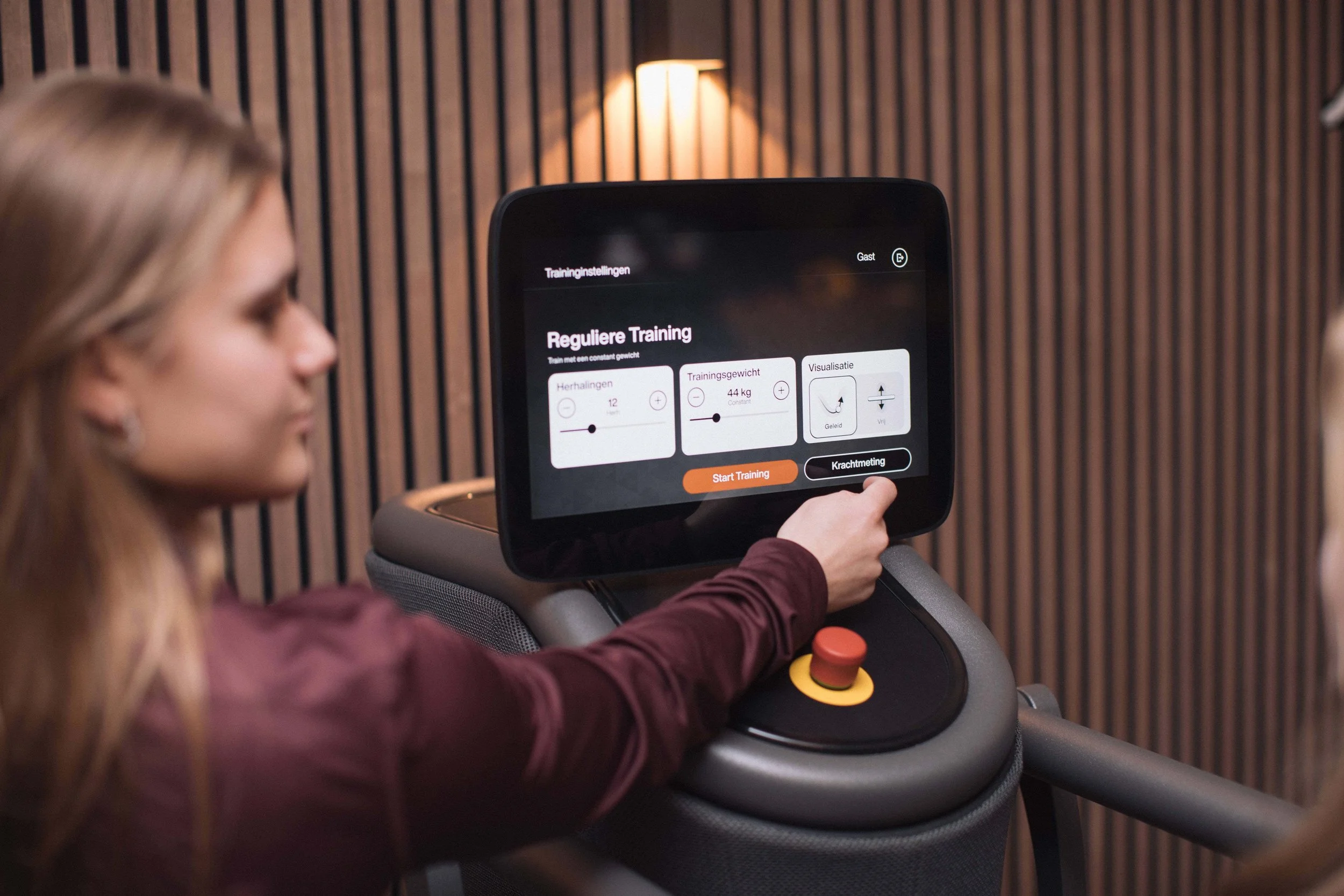 Goed Fysio Een vrouw die op een loopband traint, met een scherm dat trainingsinstellingen toont. Ze draagt een donker rode jas en de achtergrond heeft houten wandpanelen.