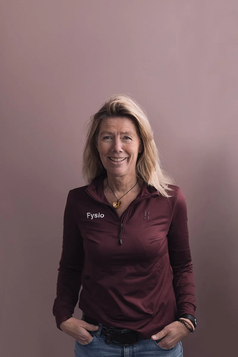 Een lachende vrouw met blond haar in een donkerrode sportzip-up en blauwe jeans, poseert tegen een effen roze achtergrond.