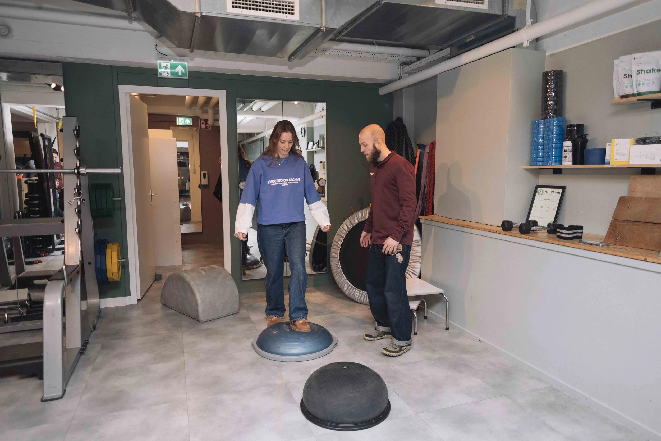 Goed Fysio Twee mensen oefenen balans op een bosu bal in een fitnessruimte, met fitnessapparatuur en wandrekken op de achtergrond.