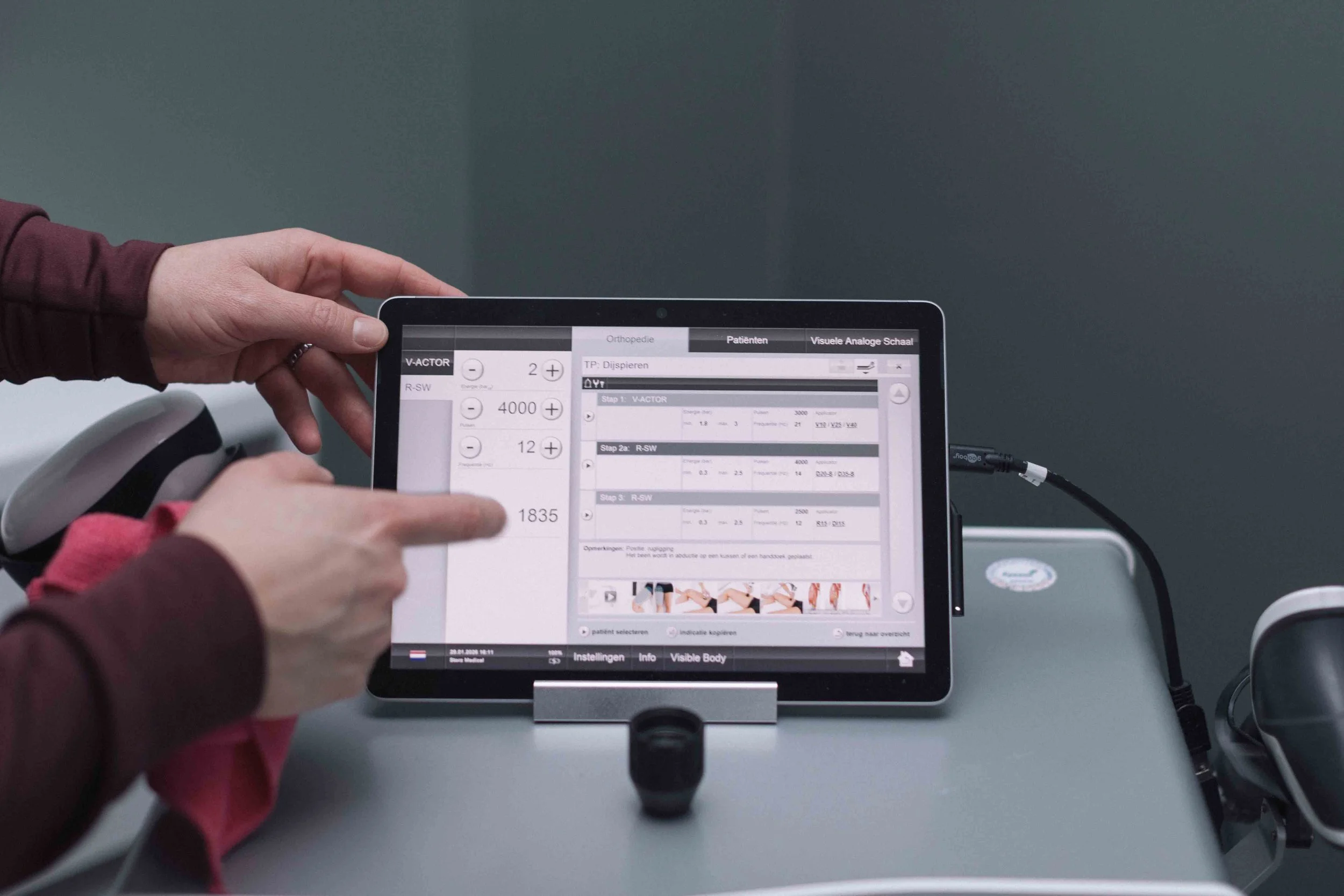 Goed Fysio Een persoon die een tablet vasthoudt met medische software die verschillende instellingen en patiëntgegevens toont, verbonden met medische apparatuur.