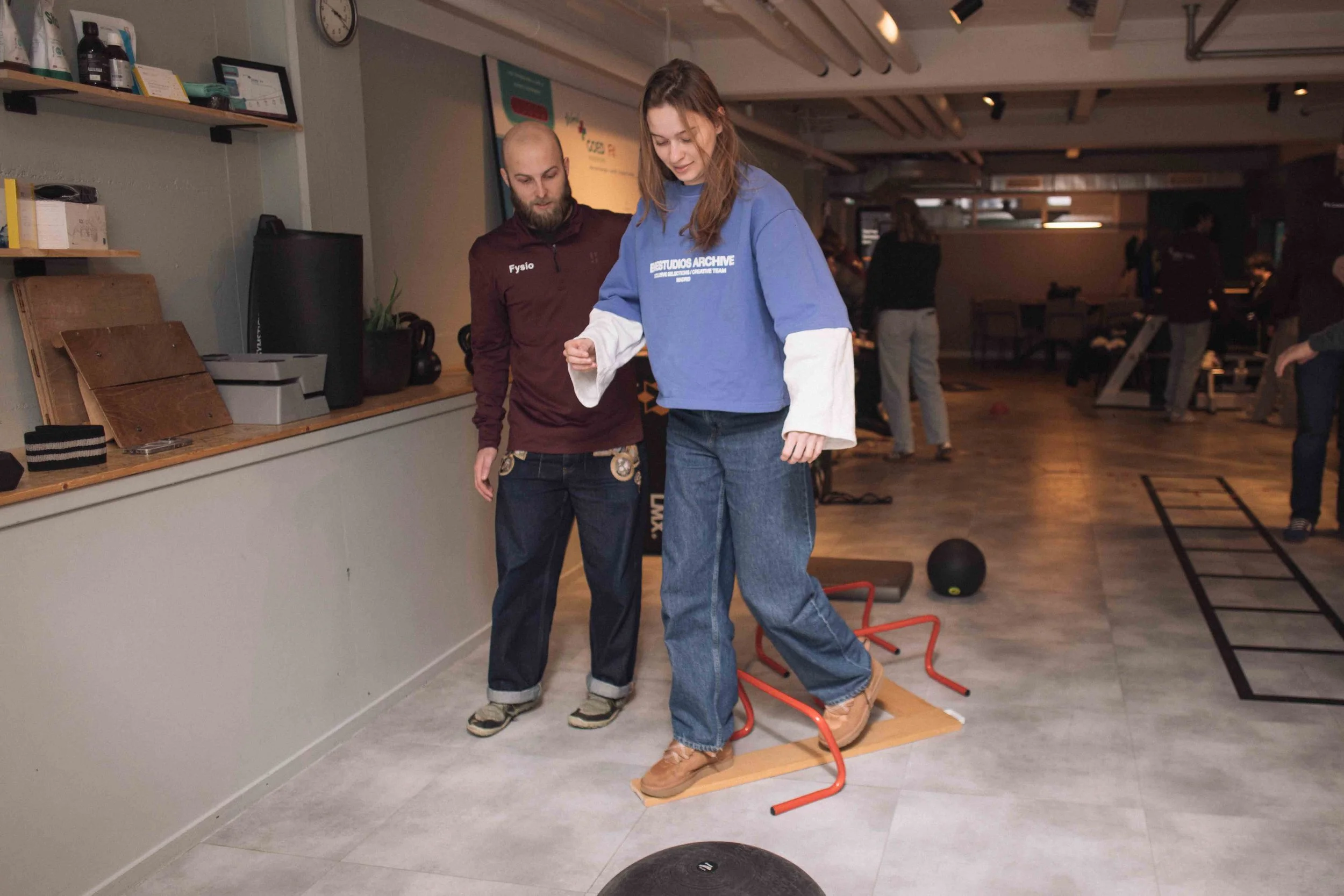 Een vrouw oefent evenwicht op een houten plank terwijl ze zich vasthoudt aan een rood-frame tijdens een groepsactiviteit in een sportschool, terwijl een instructeur haar begeleidt.