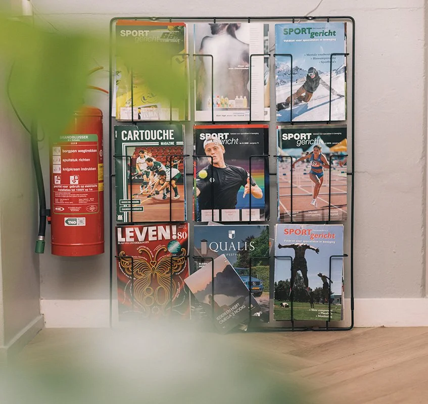 Een magazinehouder met verschillende magazines en brochures, waaronder sportmagazines en een brandblusser op een muur
