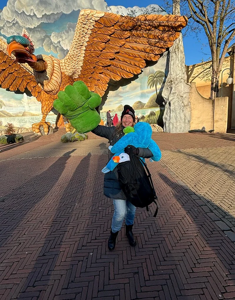Vrouw met een grote groene en blauwe plush speelgoed in een attractiepark, met een grote sculpture van een adelaar en een achtergrond met een muurschildering van een landschap.