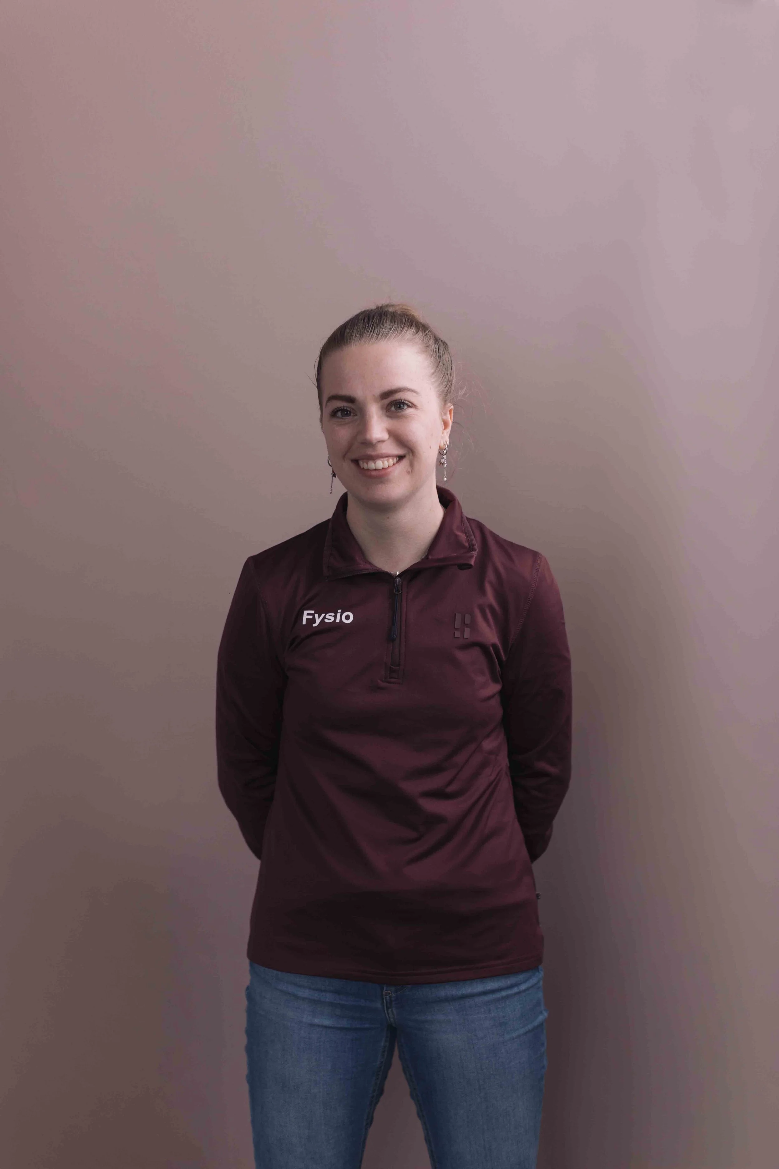 Een glimlachende jonge vrouw met haar haar in een hoge staart, draagt oorringen en een maroon sportjack met de tekst 'Fysio', staand voor een beige muur.