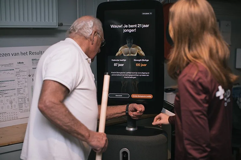 Goed Fysio Een oudere man en een jonge vrouw kijken naar een groot digitaal scherm dat een transhumanistische app toont met een bericht dat iemand 21 jaar jonger is, specifiek 66 jaar in plaats van 87 jaar.