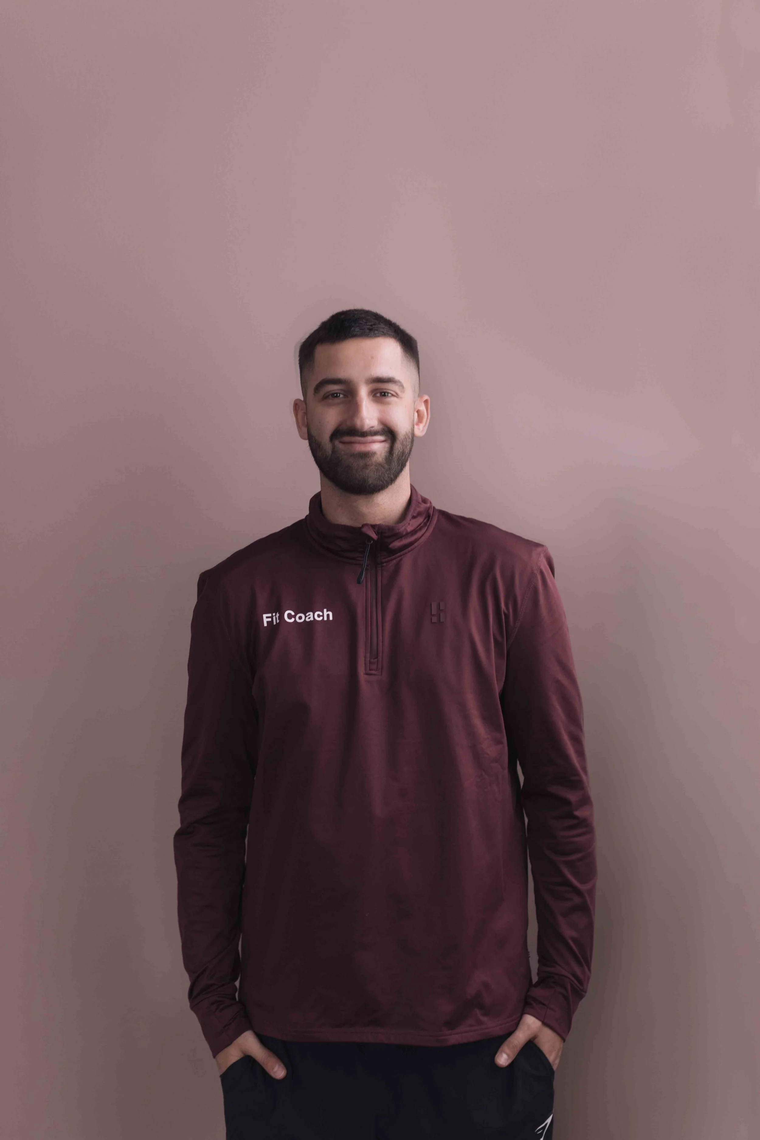 Michael Hemmes Goed Fysio Man met een baard en korte haren, lachend, draagt een maroon trainingsjack met de tekst 'Fit Coach' en een logo, staand voor een lichtbruine muur.