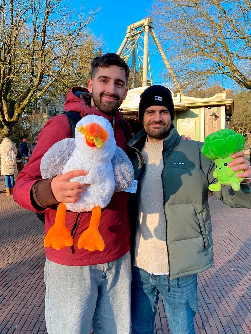 Twee mannen in een pretpark, één houdt een grote pluchen eend vast en de andere een pluchen broccoli, beiden glimlachen.