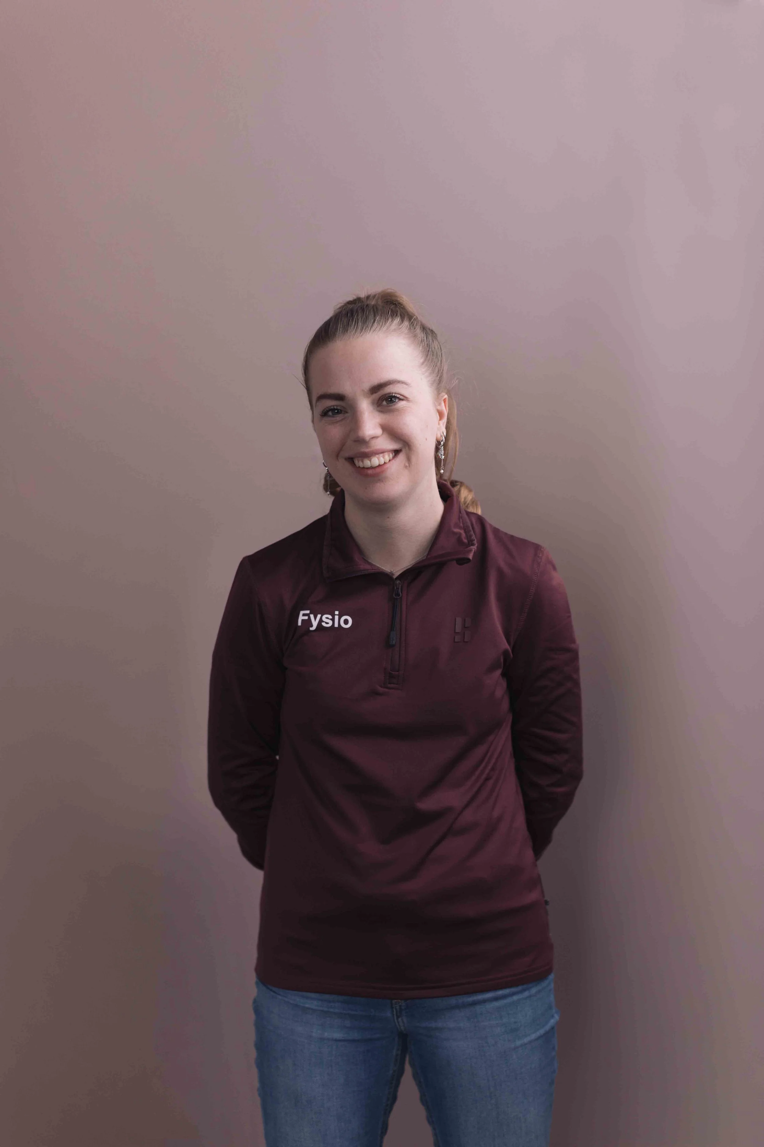 Sophie Pigmans Goed Fysio een vrouw met een glimlach, gekleed in een donkerrode sportjas met de tekst 'Fysio' op de linkerzijde, en blauwe jeans, staat voor een effen lichtpaarse muur.