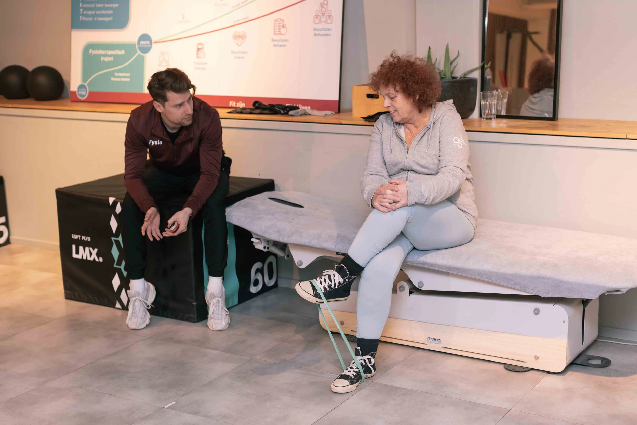 Goed Fysio Een man in sportkleding die zich voorbereidt op een oefening met een band rondom zijn enkel, terwijl hij naast een vrouw op een medische behandeltafel zit in een sportschool of fysiotherapiepraktijk.
