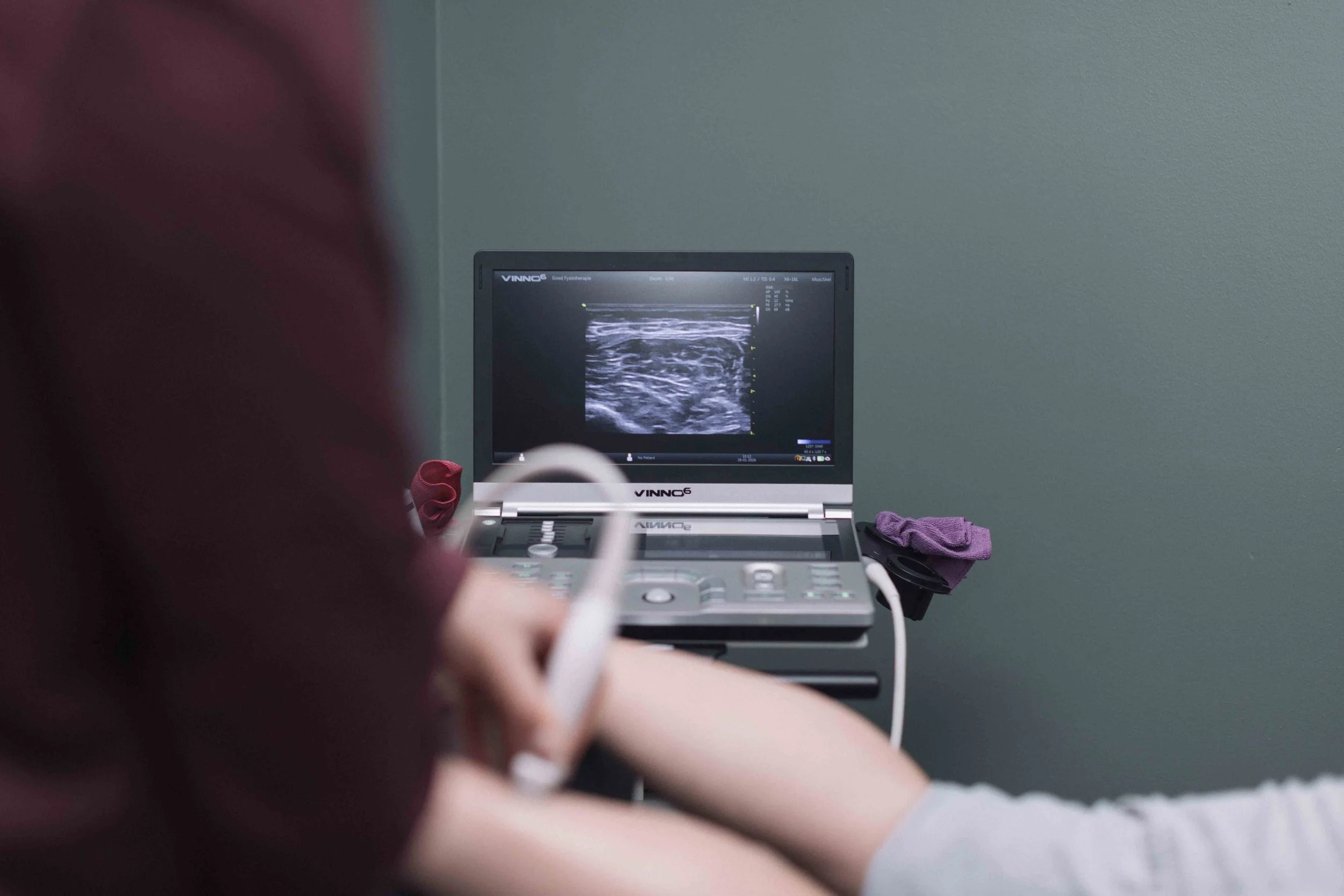 Goed Fysio Een persoon gebruikt een draagbare echografie-apparaat in een medische setting, met een monitor die een echografiebeeld toont.