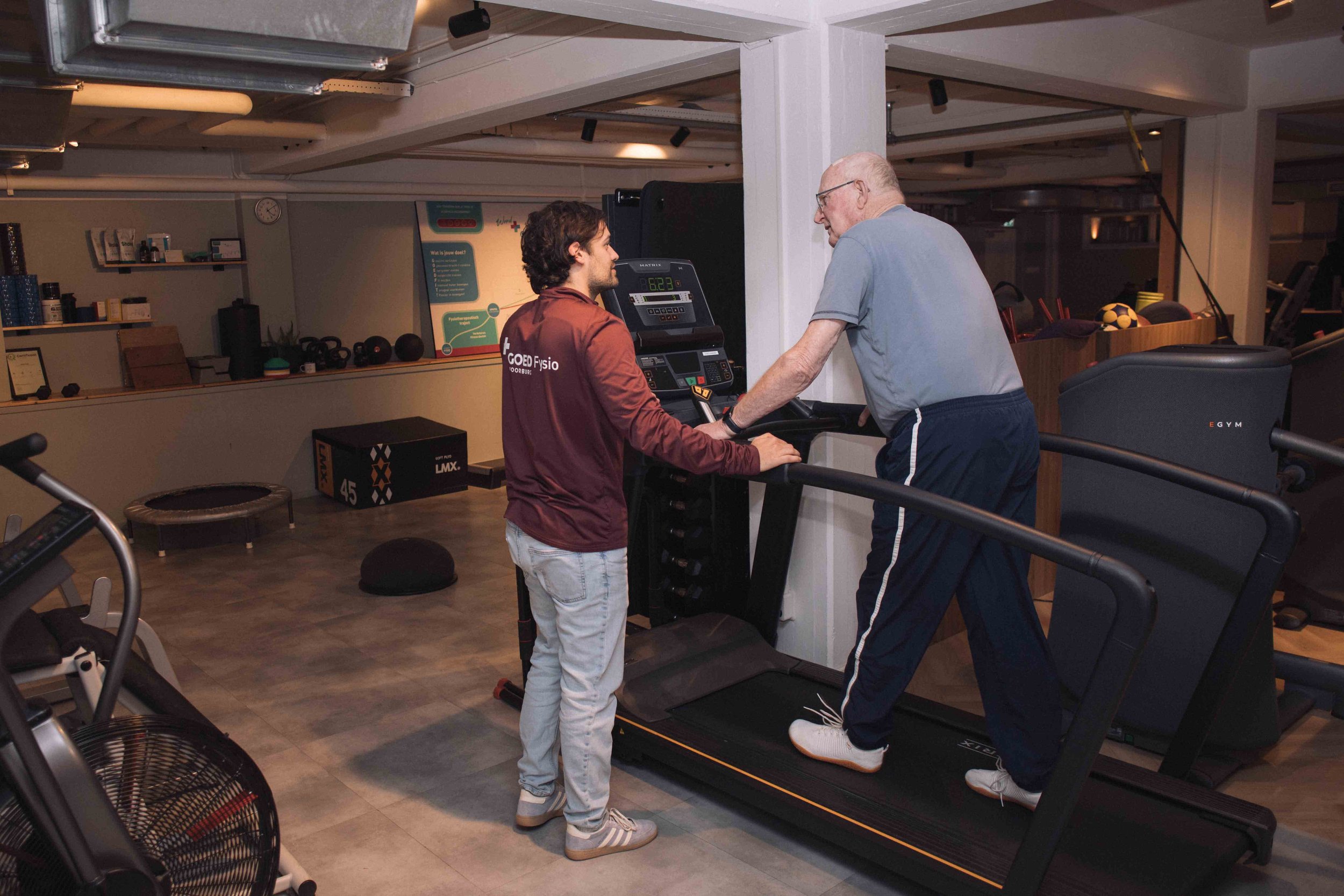 Goed Fysio Een jonge man en een oudere man trainen samen op een loopband in een sportschool.