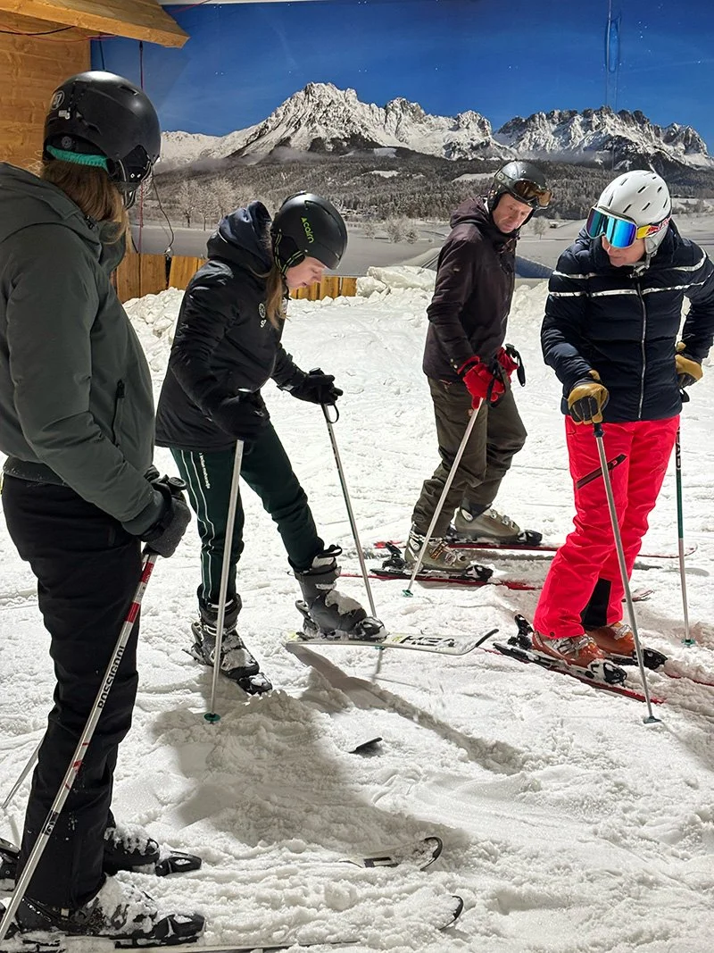 Vijf mensen leren skiën op een skibos, met een berg en besneeuwd landschap op de achtergrond.