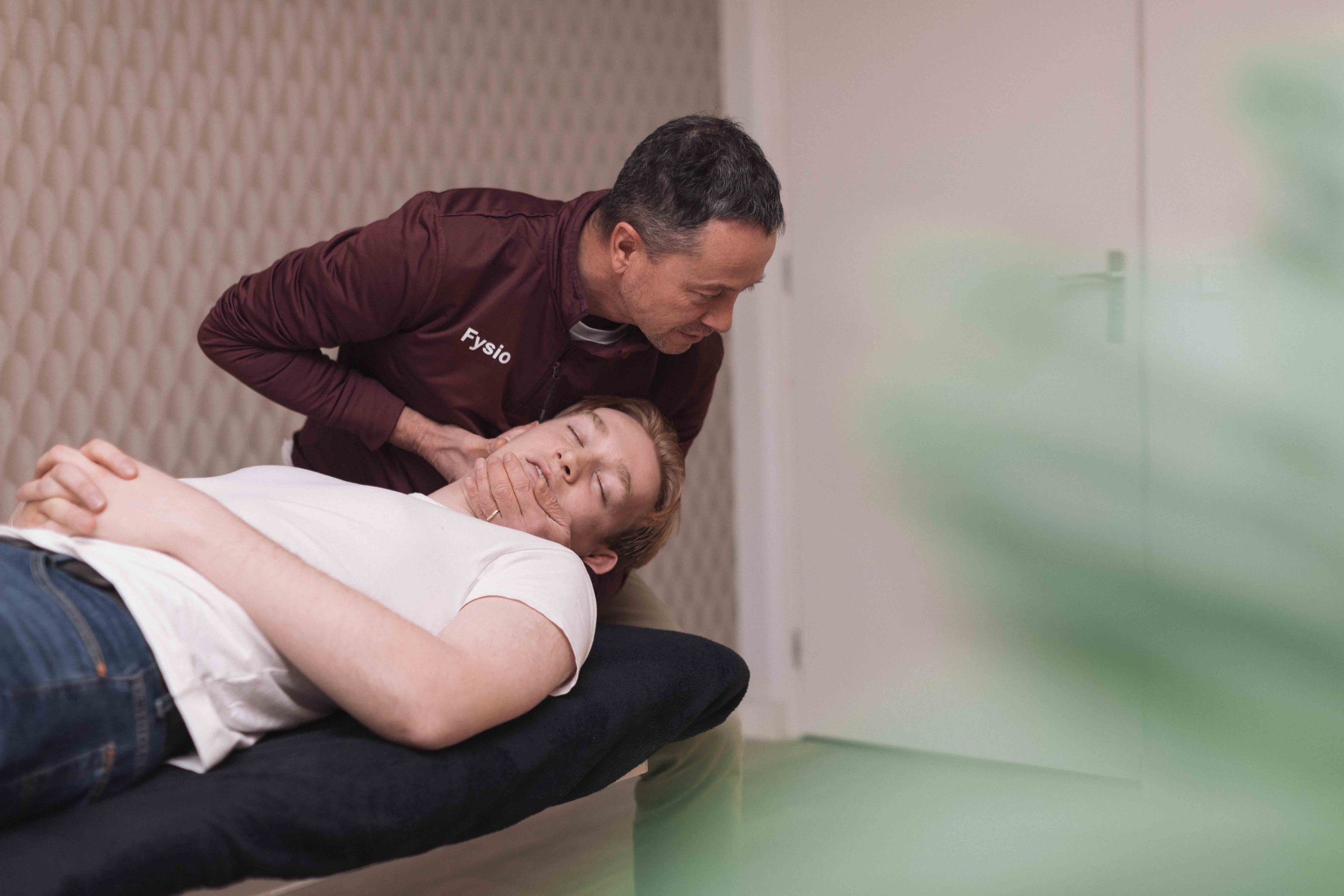 Goed Fysio Een man in een donkerrode jas met het woord 'Fysio' erop, die een vrouw reanimeert op een bed in een medische omgeving.