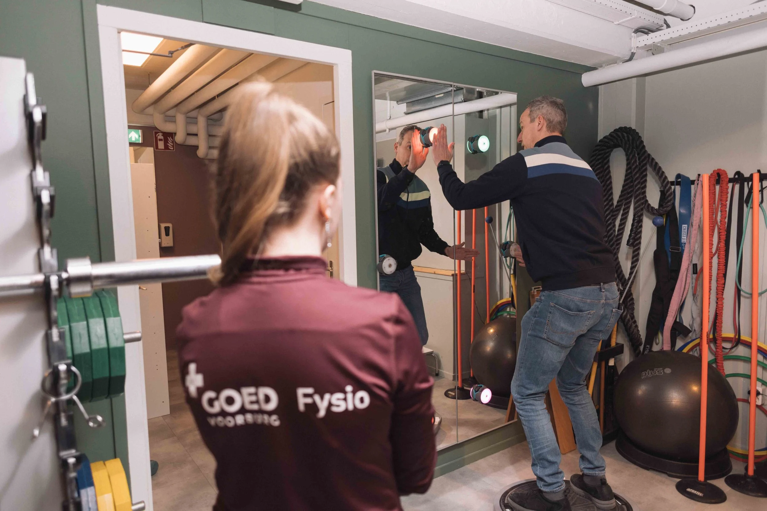 Goed Fysio Twee mensen oefenen balans op een draaiende skate plaat terwijl een vrouw toekijkt, in een sportschool met verschillende fitnessapparatuur aan de muur en een grote spiegel.