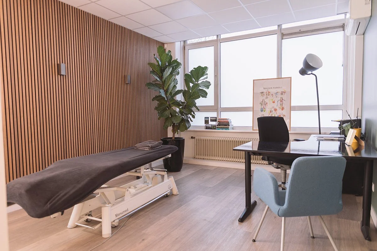 Een kantoor met een bureau, een computerscherm, een bureaustoel, een lamp, een muur met houten lamellen, een grote plant, een rugzak, een behandeltafel en een raam met frosted glas.