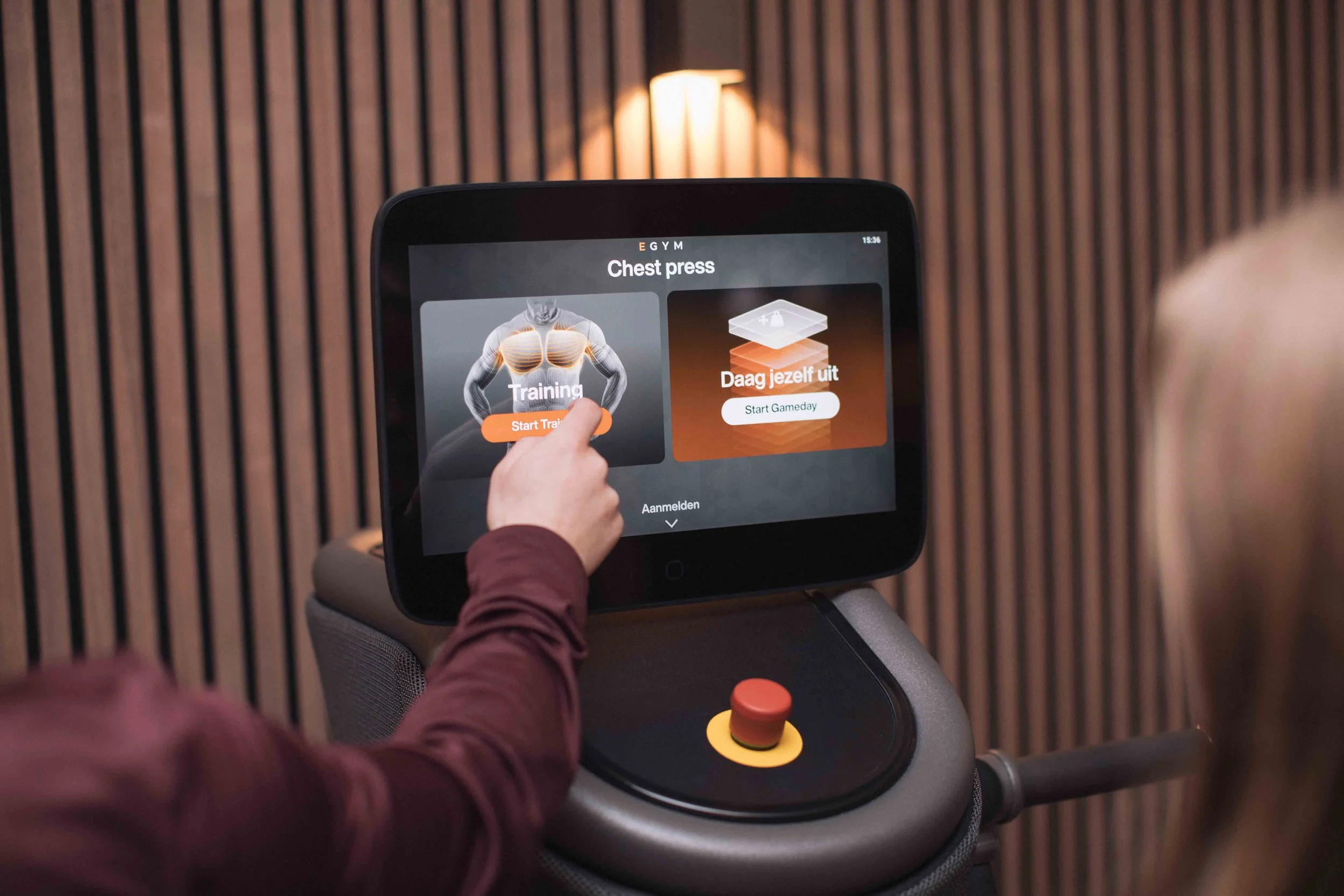 Goed Fysio Een persoon bedient een fitnessapparaat met een touchscreen dat opties voor training en ontspanning toont, in een sportschool met houten wandpanelen.