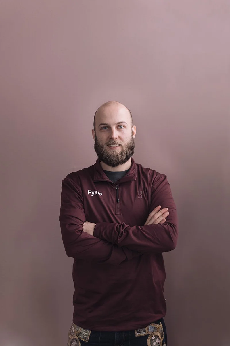 Tim Oorschot Goed Fysio Man met een baard, gekleed in een paarse sportjack met rits en tekst 'Fysio' op de linkerkant, met armen over elkaar, staat voor een effen, lichtroze achtergrond.