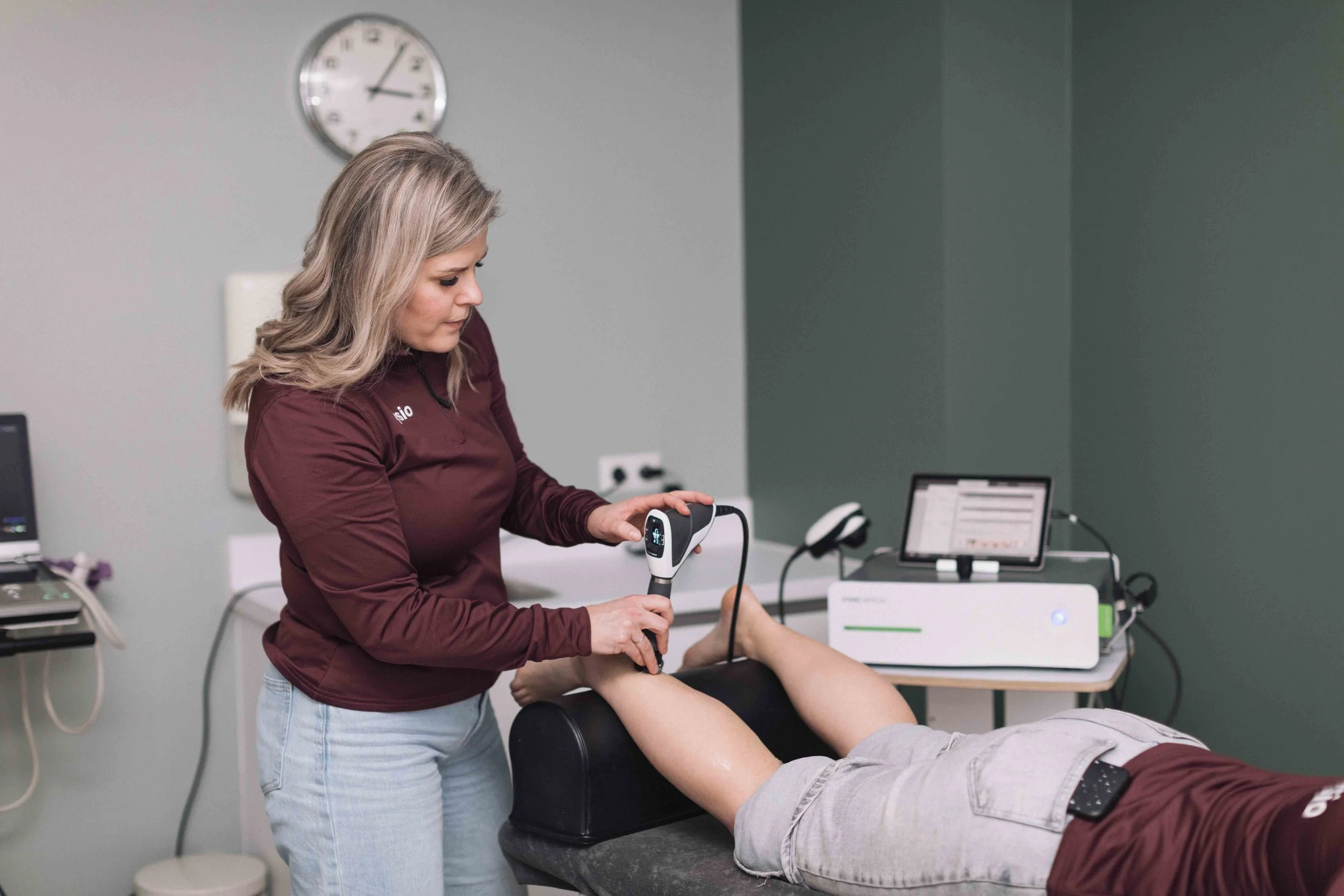 Goed Fysio Een vrouw onderzoekt een arm tijdens een medische test of behandeling in een kliniek, met medische apparatuur op een tafel en een laptop op de achtergrond.