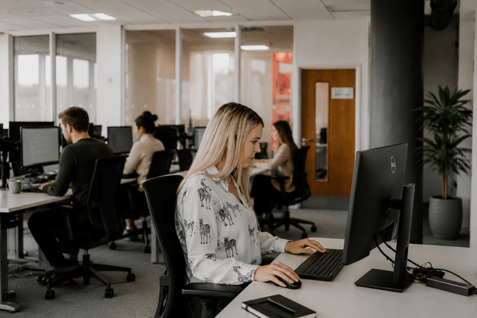 Woman working at a desk in an office with other employees working at computers in the background.