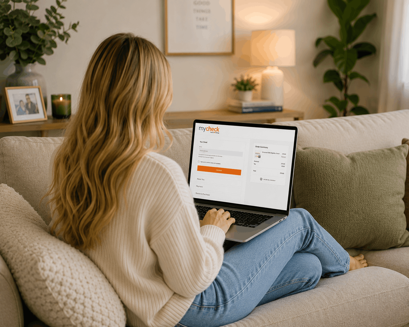 Woman sitting on a beige sofa using a laptop to check an order on a website called mycheck, in a cozy living room with a framed photo, candle, and lamp in the background.