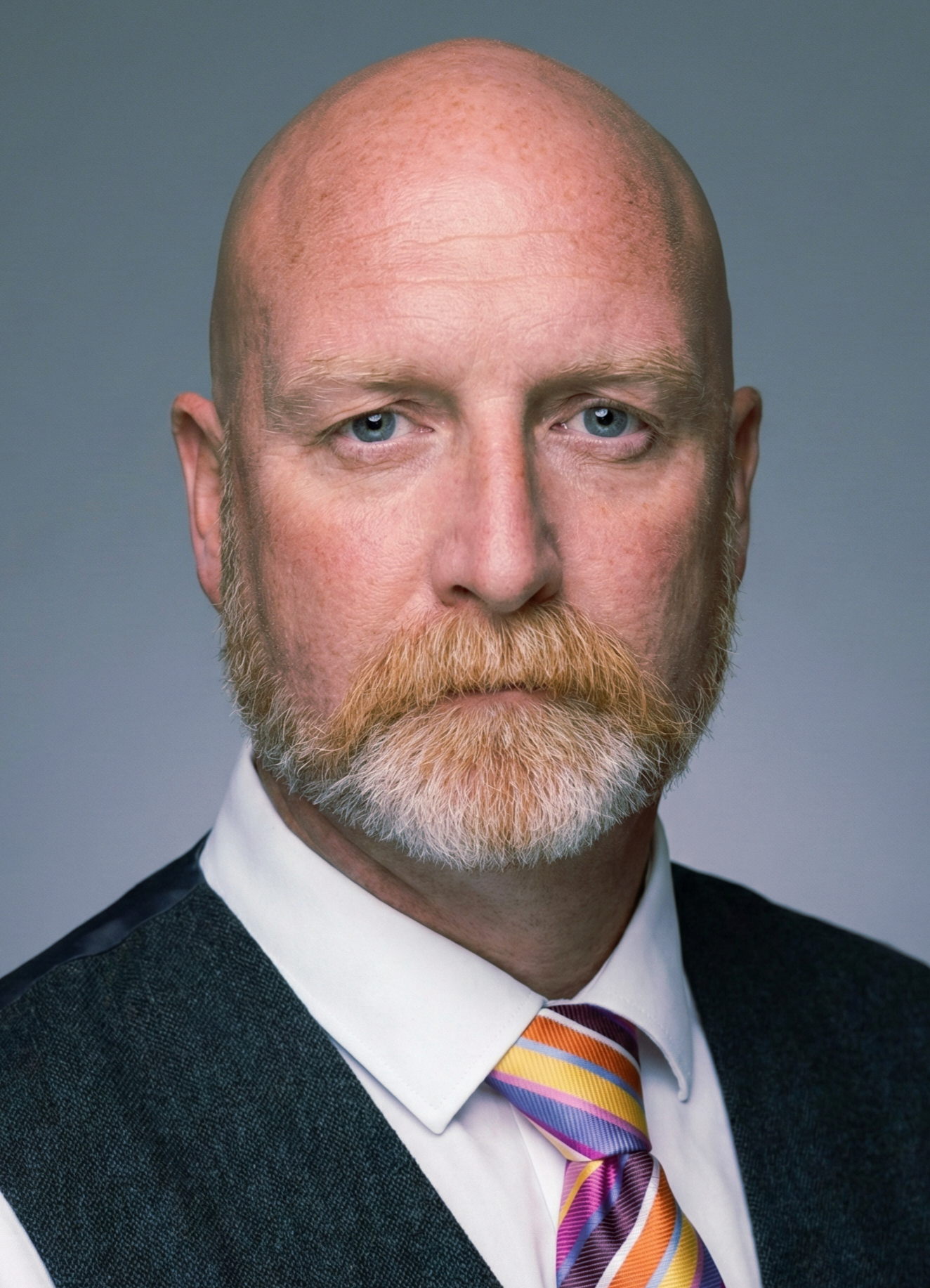 Portrait d'un homme avec une barbe et une tête rasée, portant une veste, une chemise blanche et une cravate rayée multicolore.
