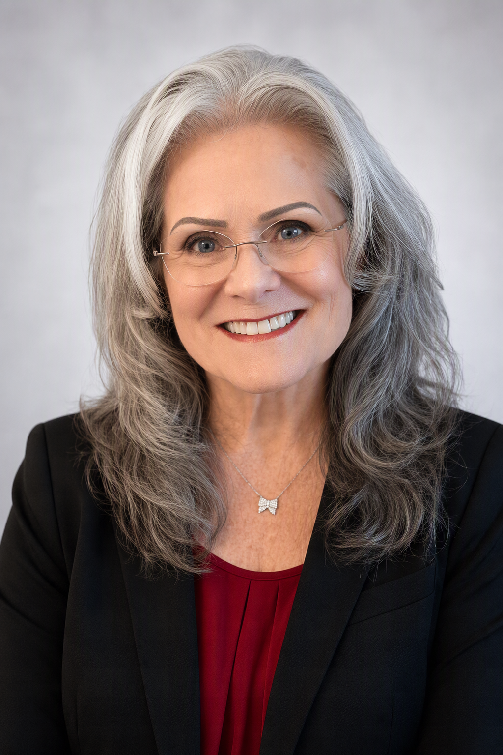A smiling middle-aged woman with long gray hair, wearing glasses, a black blazer, and a red top. She has a butterfly necklace and is looking directly at the camera.