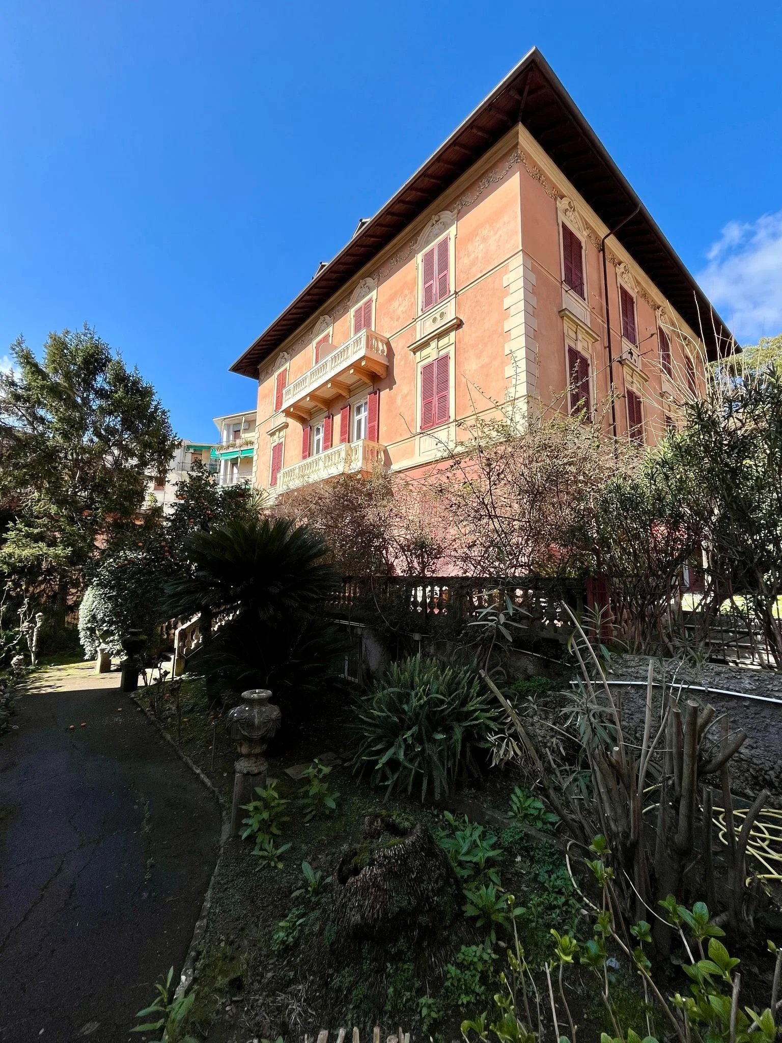 Un edificio storico di colore rosa con finestre con tapparelle rosse, un balcone e una tettoia di legno, circondato da un giardino con piante e arbusti, sotto un cielo blu con alcune nuvole.