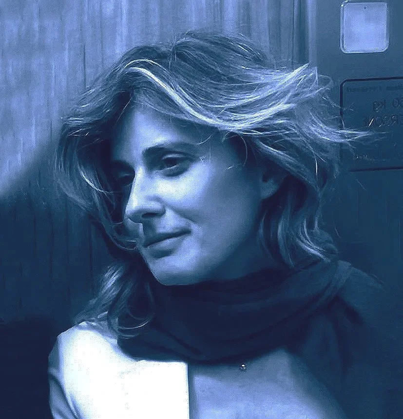 Ritratto di una donna con capelli mossi e risvolti bianchi, che sorride leggermente, fotografata con effetto monocromatico blu in un ambiente con pareti di legno.
