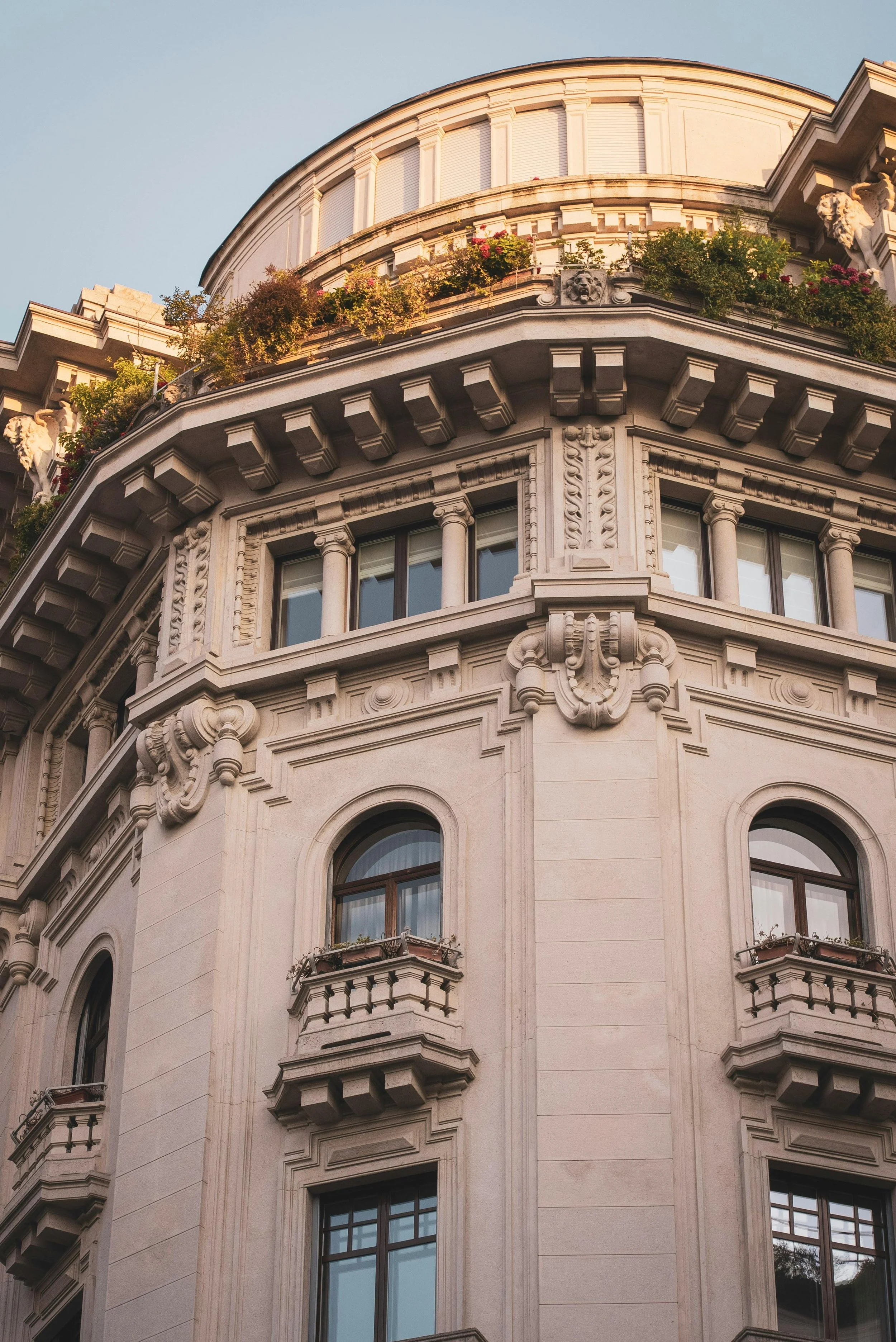 Edificio in stile classico con dettagli decorativi in pietra, balconi con ringhiera e piante sui davanzali, al tramonto.