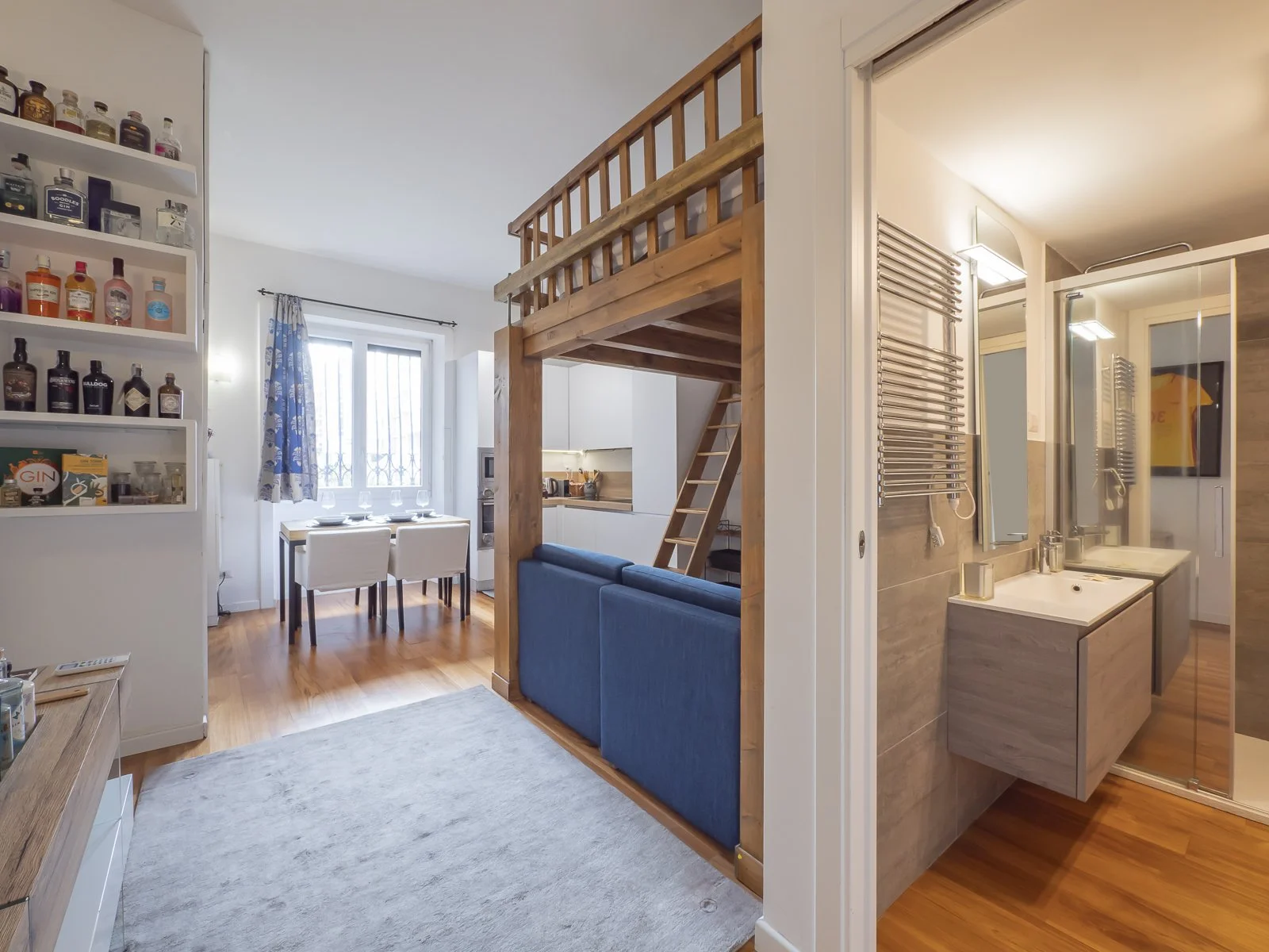 Interno di un appartamento con pareti bianche, pavimento in legno, zona soggiorno con divano blu, cucina con zona pranzo con tavolo e sedie, e una scala in legno che porta a un soppalco. Bash in bagno con lavabo e specchio.