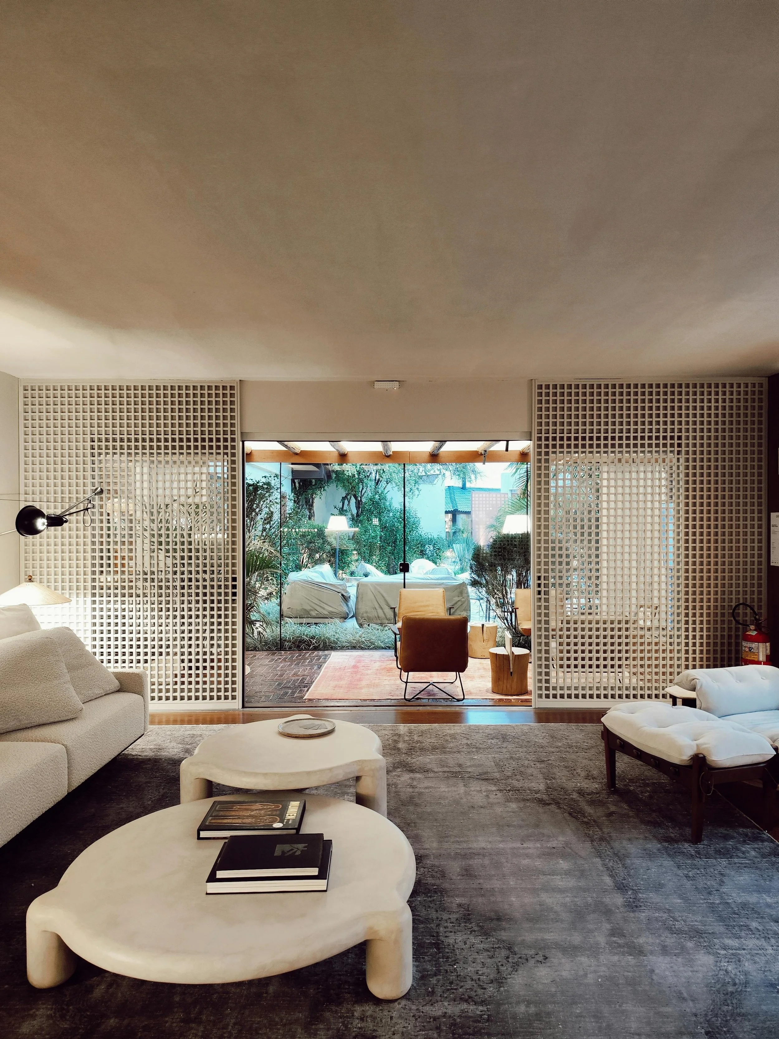 Interno di un soggiorno elegante con divano beige, tavolini di vetro, poltrona di pelle e pareti divisorie in legno a griglia, con una vista su un giardino esterno con sedie e tavoli.