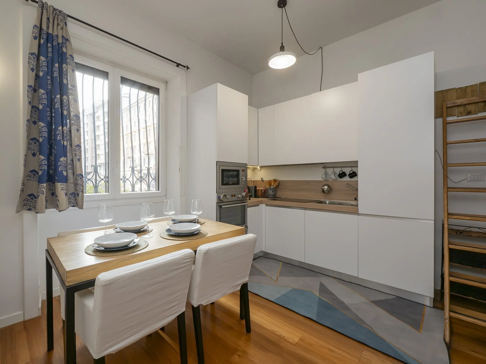 Cucina moderna con tavolo da pranzo, dotata di piatti e bicchieri, finestra con tende Blu e Beige, armadi bianchi, lavello e parete di legno, scaffale in legno.