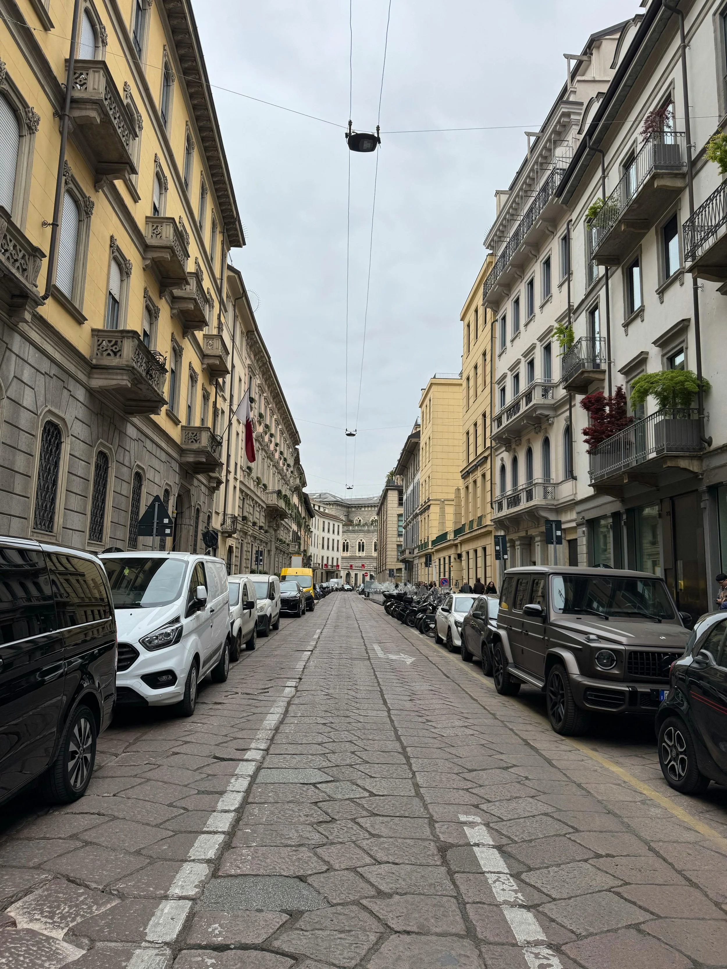 Via Urbana con edifici storici e negozi ai lati, auto parcheggiate lungo i marciapiedi e cielo coperto