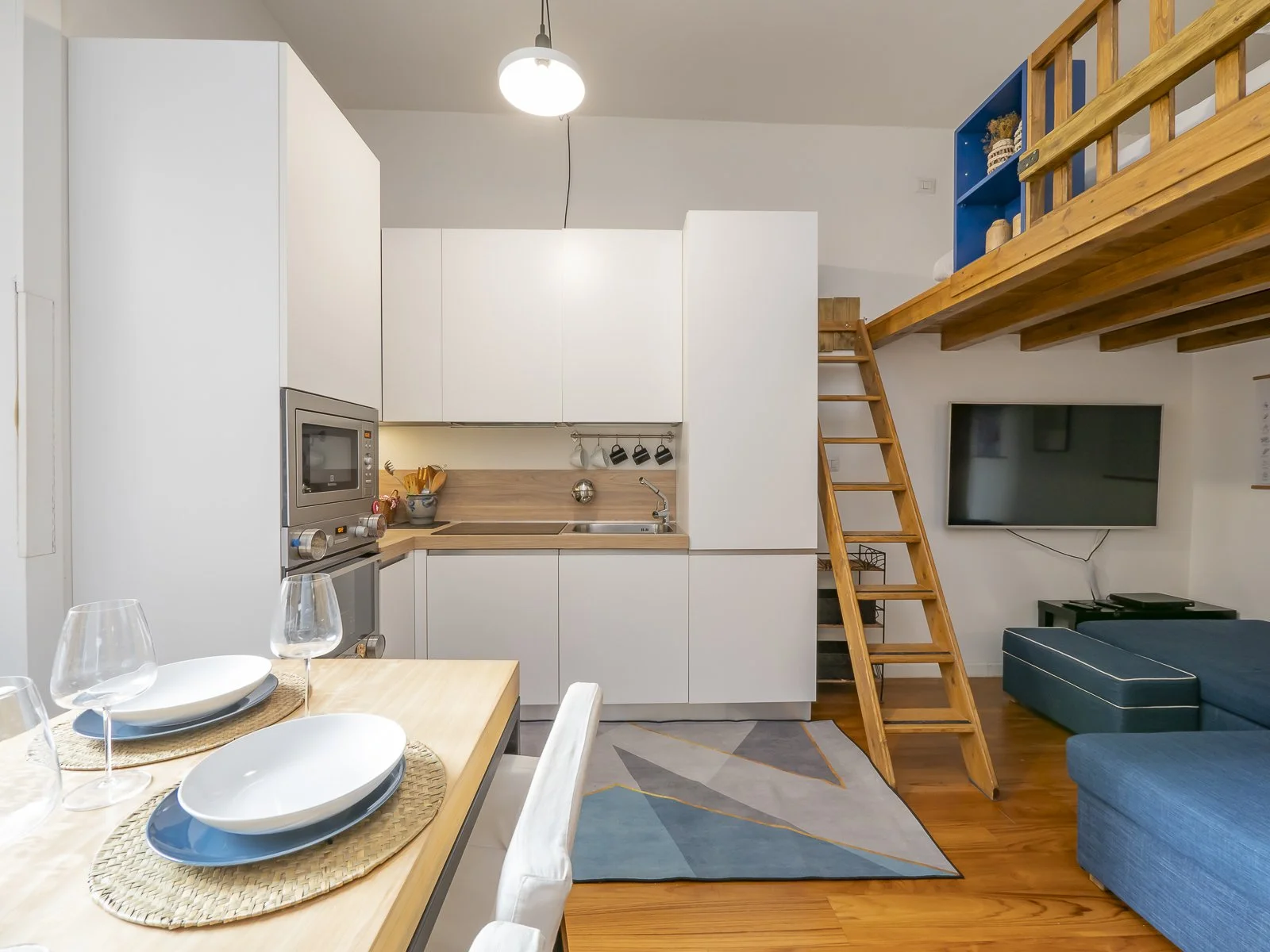 Interno di un appartamento, vista dalla zona pranzo verso la cucina e il soggiorno, con un soppalco e una scala in legno.