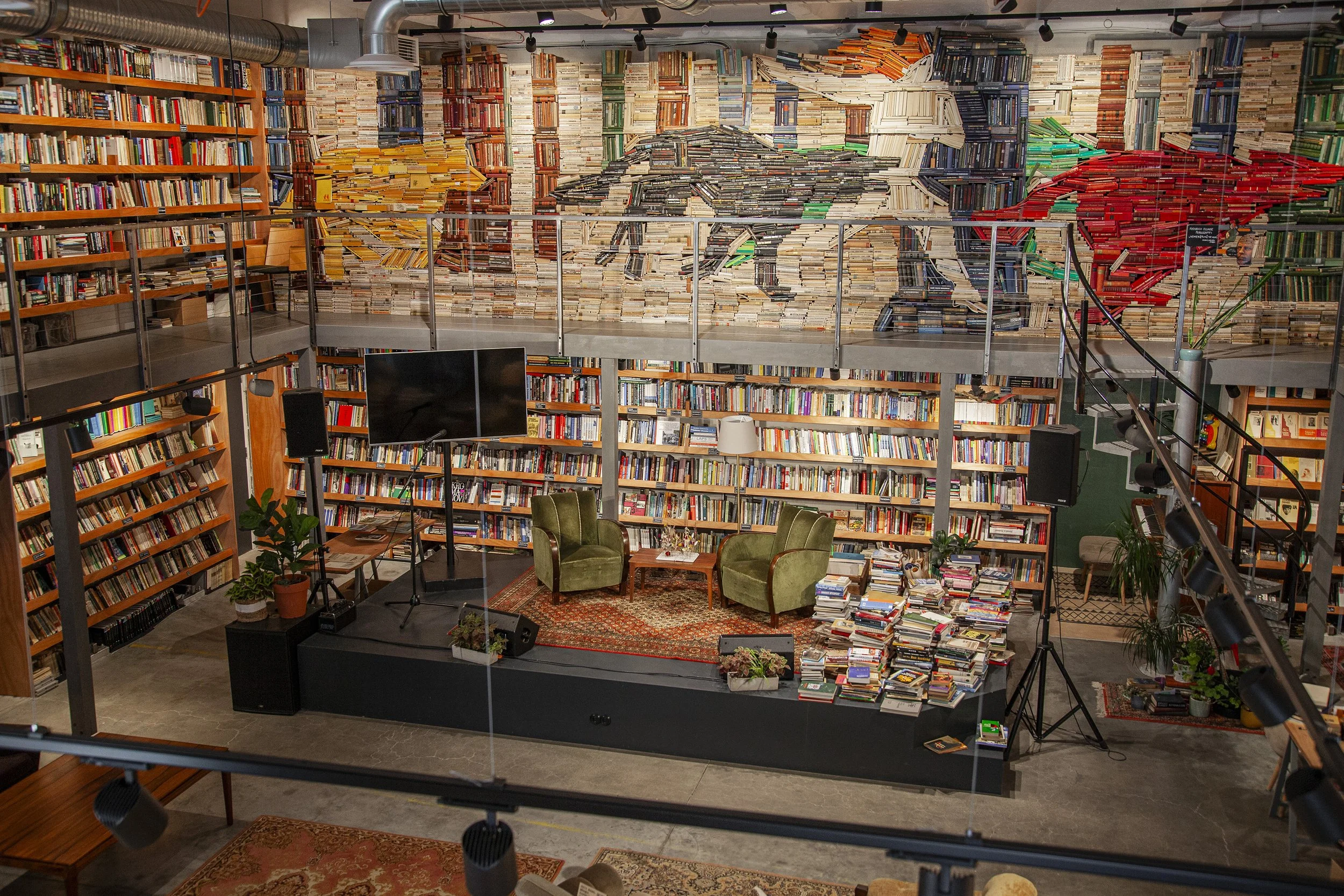 Interior of a bookstore or library with a small stage setup for a talk or presentation, surrounded by numerous bookshelves and a wall mural of a horse painted with books. There are stacks of books on the floor, a few potted plants, green armchairs, a