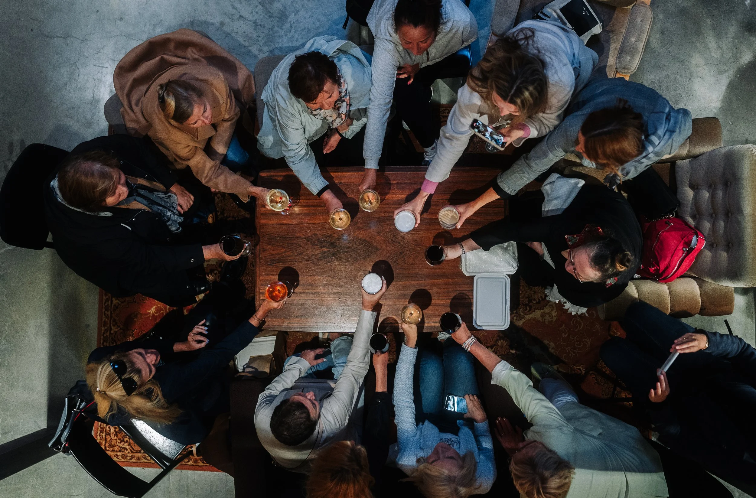 Overhead view of a group of people gathering around a wooden table, raising glasses with drinks to toast, in an indoor setting.