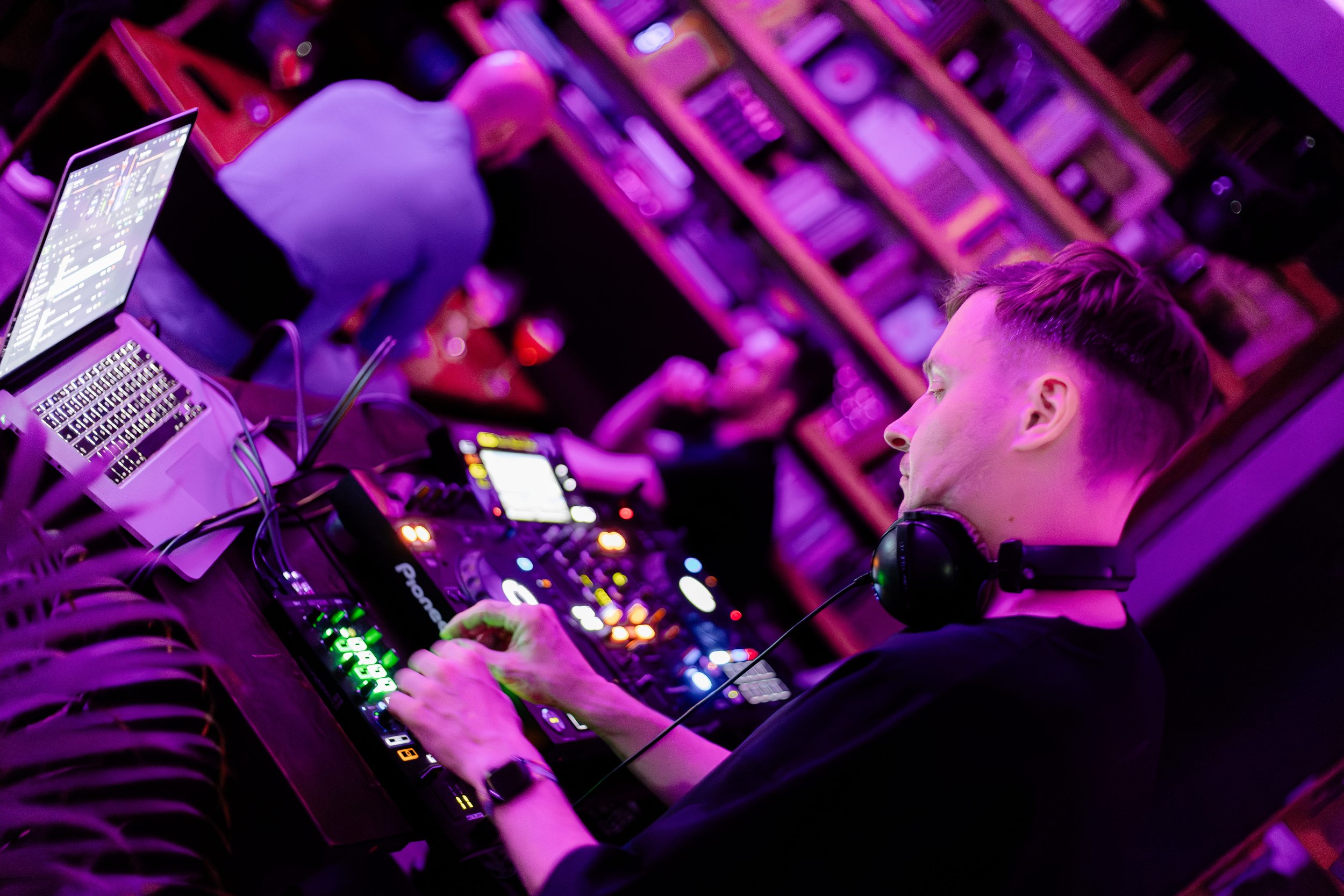 DJ wearing headphones operating electronic music equipment in a nightclub with purple lighting