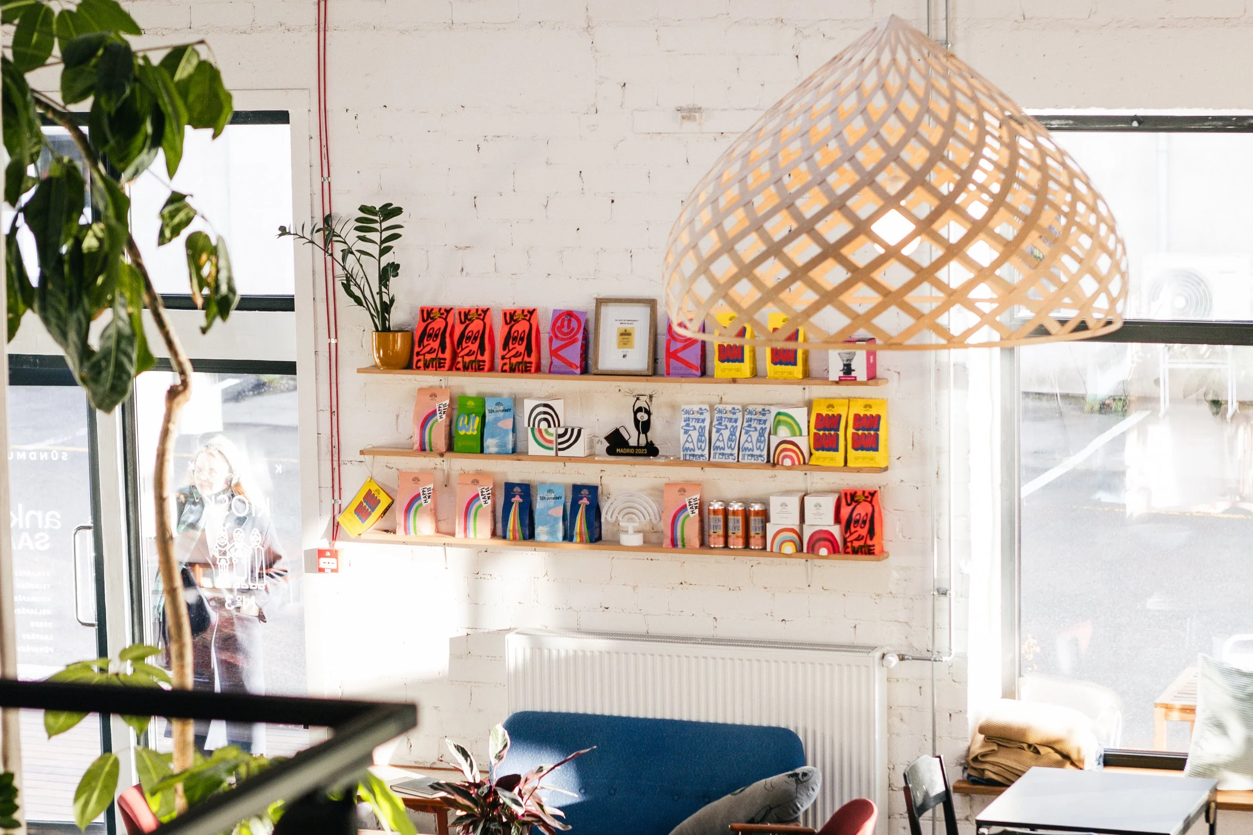Interior of a store or cafe with shelves displaying colorful books and decorative items, a large woven pendant light, and a person visible outside through glass door.