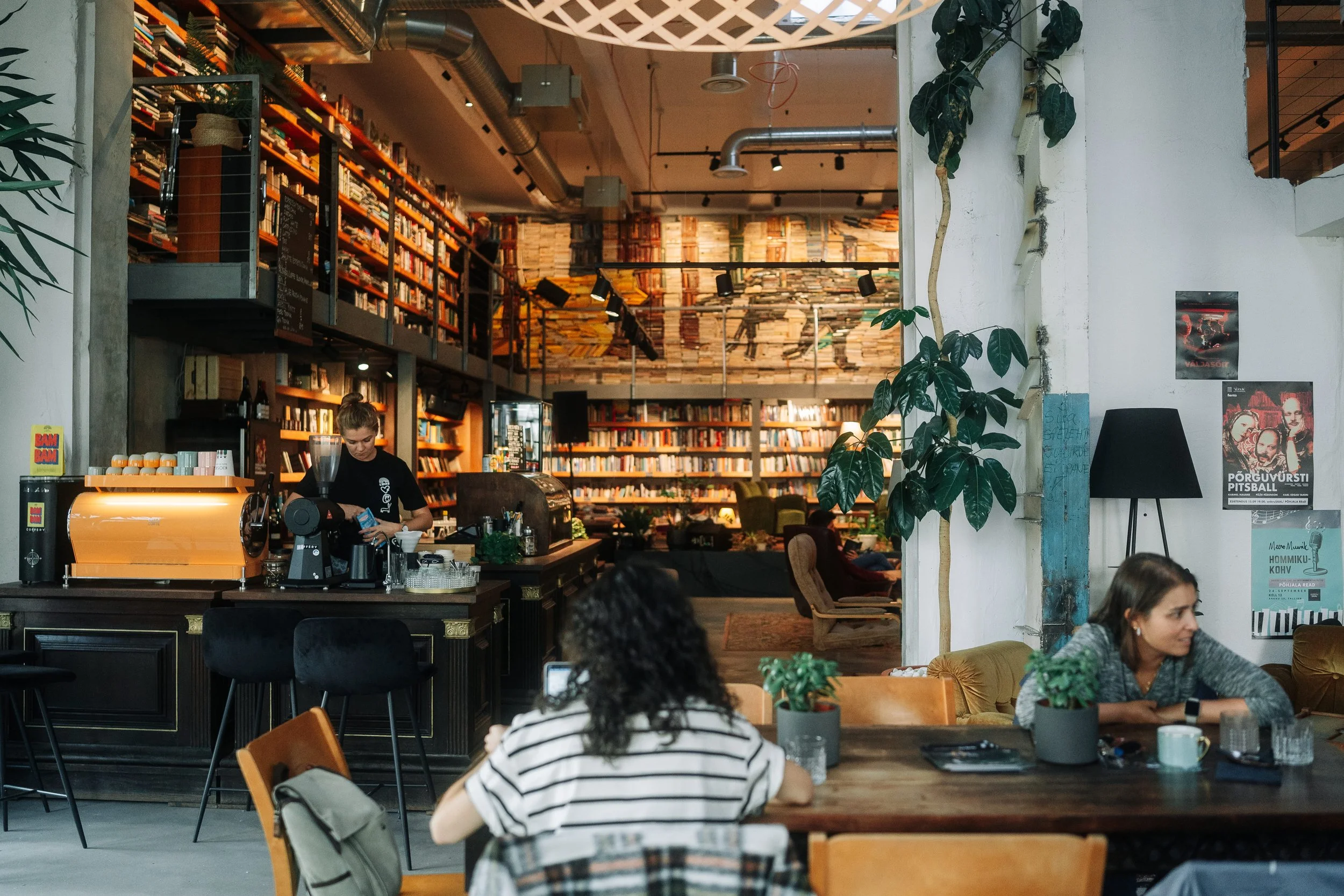 Interior of a cozy coffee shop with a bookshelf-filled wall, a coffee counter with an espresso machine, and customers sitting at tables, some working on laptops.