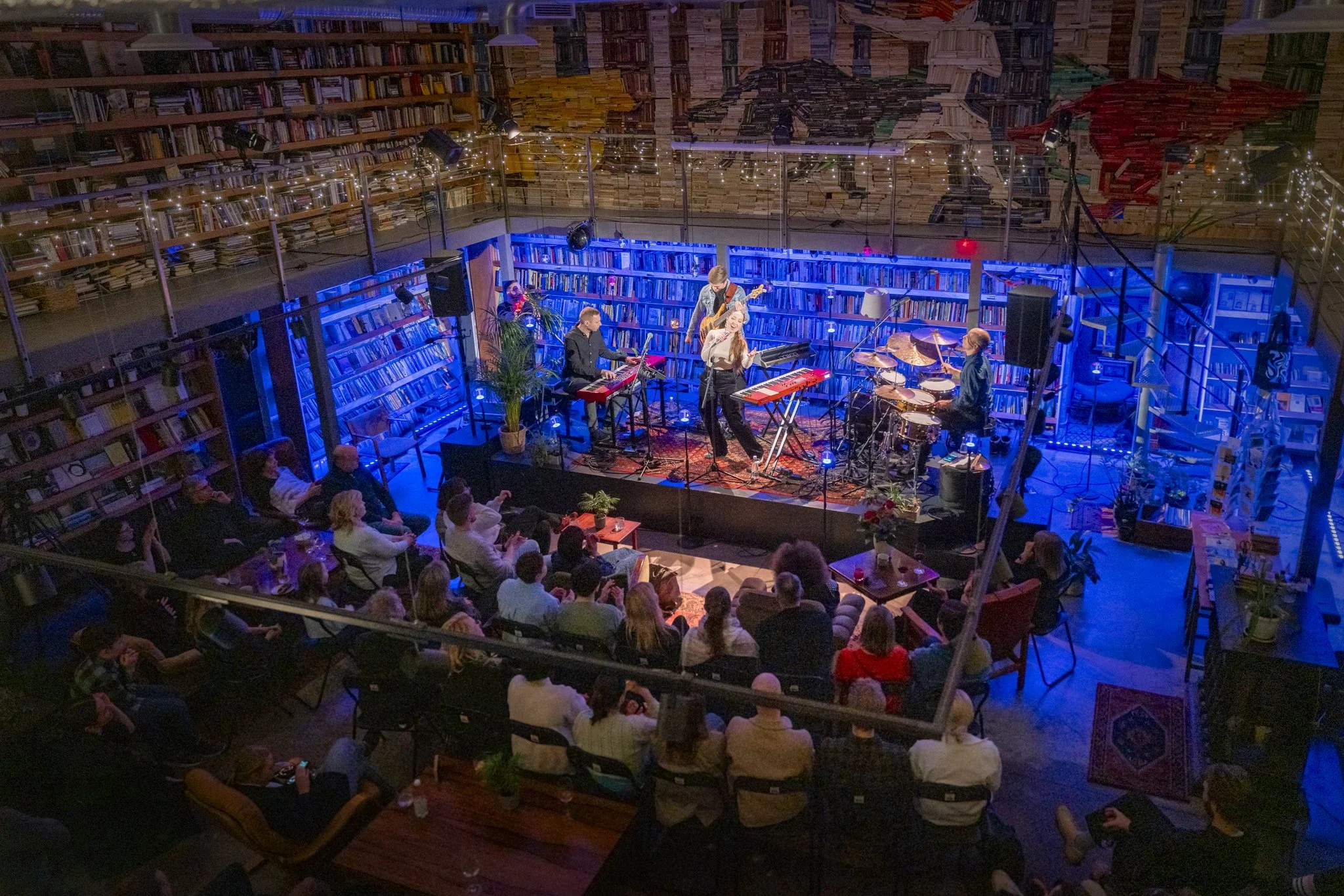 A live music performance in a cozy, multi-level venue with an audience seated around the stage. The stage features a band with instruments including keyboards, guitar, drums, and a singer with a microphone. The background is filled with bookshelves, 