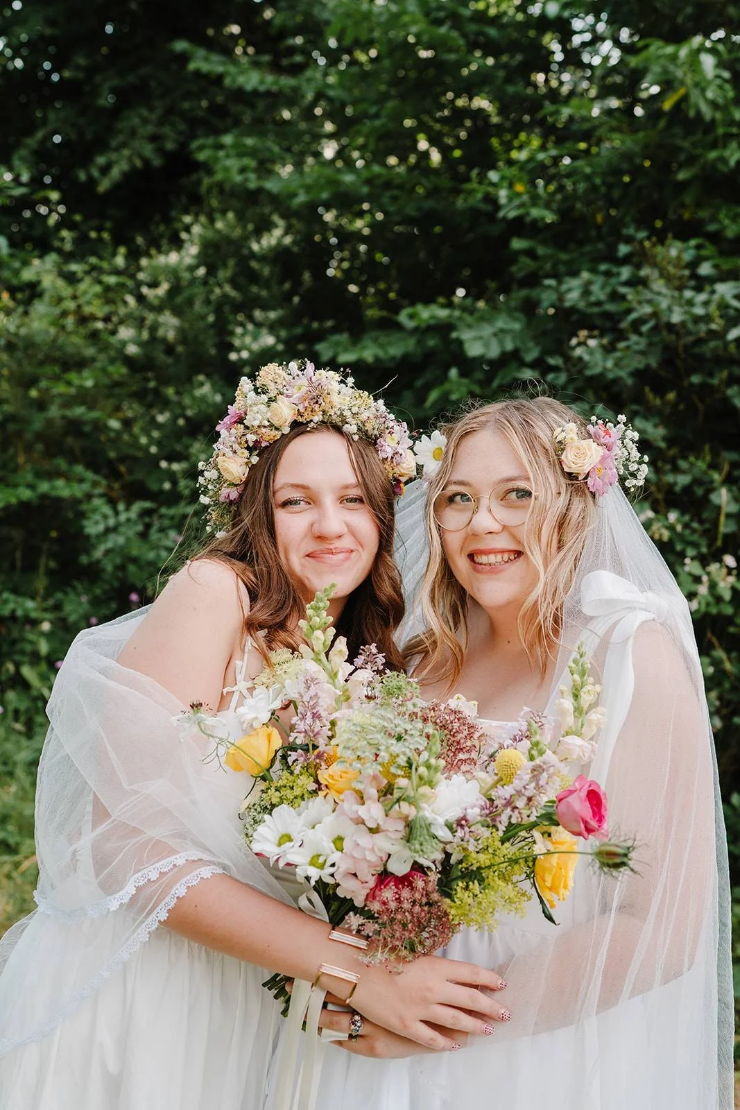 Two women in wedding dresses holding a bouquet of flowers, wearing flower crowns, outdoors with green foliage background.