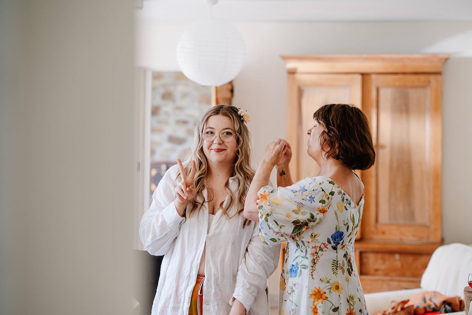 A young woman with glasses and a flower in her hair making a peace sign while smiling and standing next to an older woman holding her hand, in a bright room with a wooden cabinet and hanging paper lanterns.