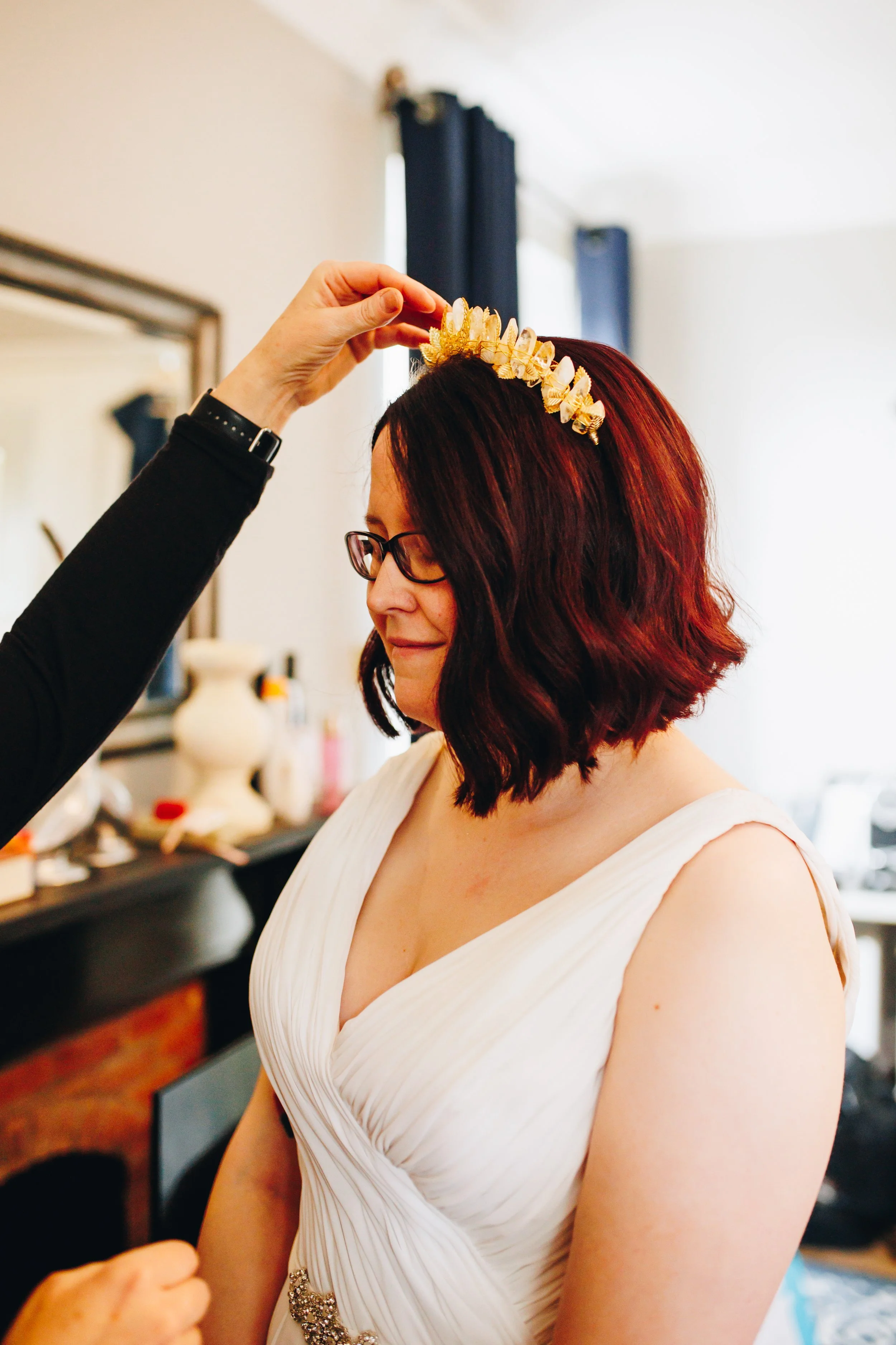 A woman with shoulder-length dark hair, wearing glasses and a white dress, is smiling softly as someone places a gold crown or tiara on her head in a room with a mirror and blue curtains.