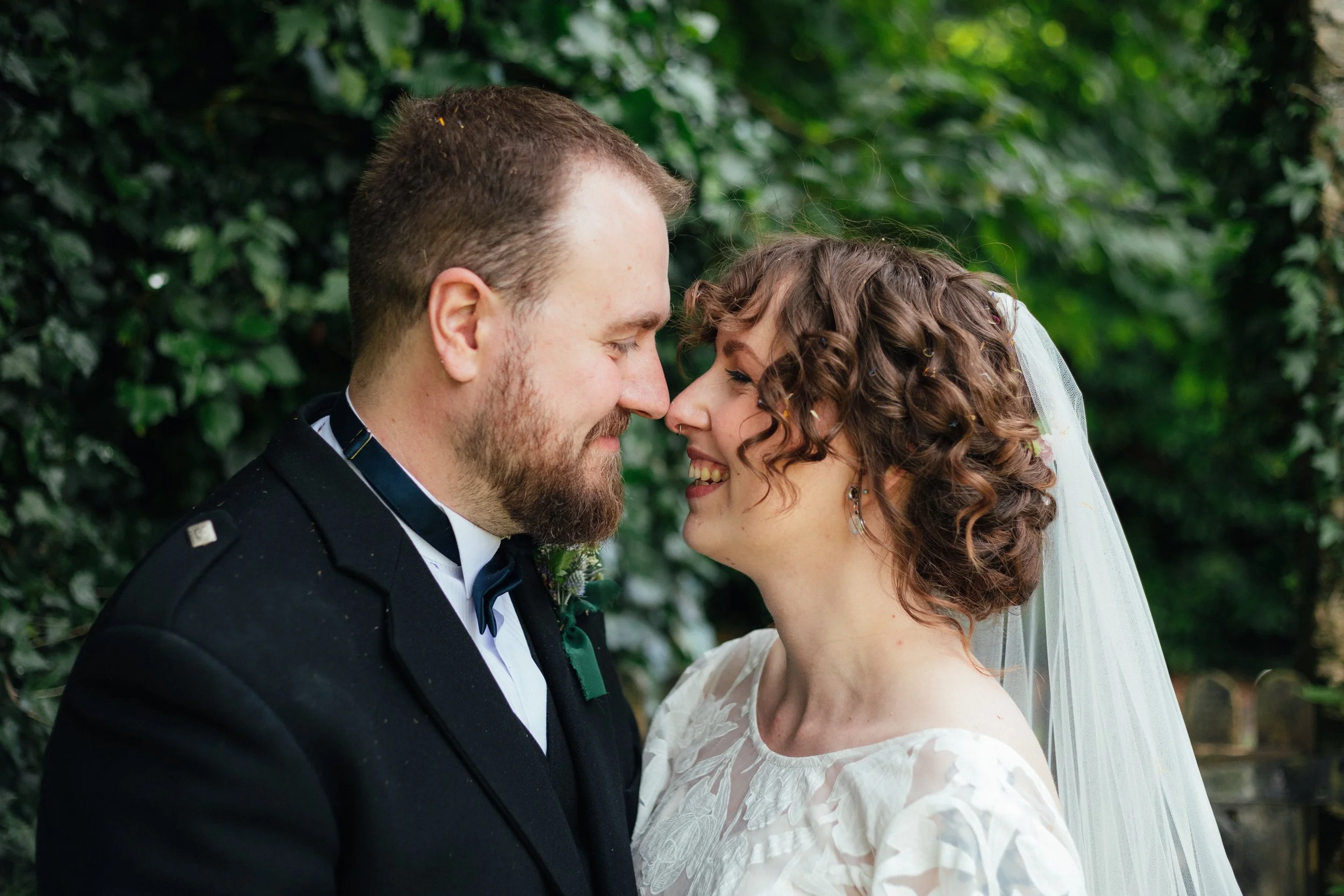 A bride and groom facing each other closely on their wedding day, surrounded by greenery.