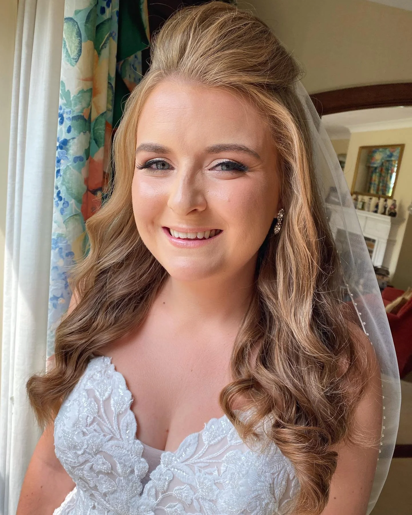 Close-up of a smiling bride with wavy hair, wearing a white lace wedding dress and a veil, standing near a window with floral curtains in a warmly lit room.
