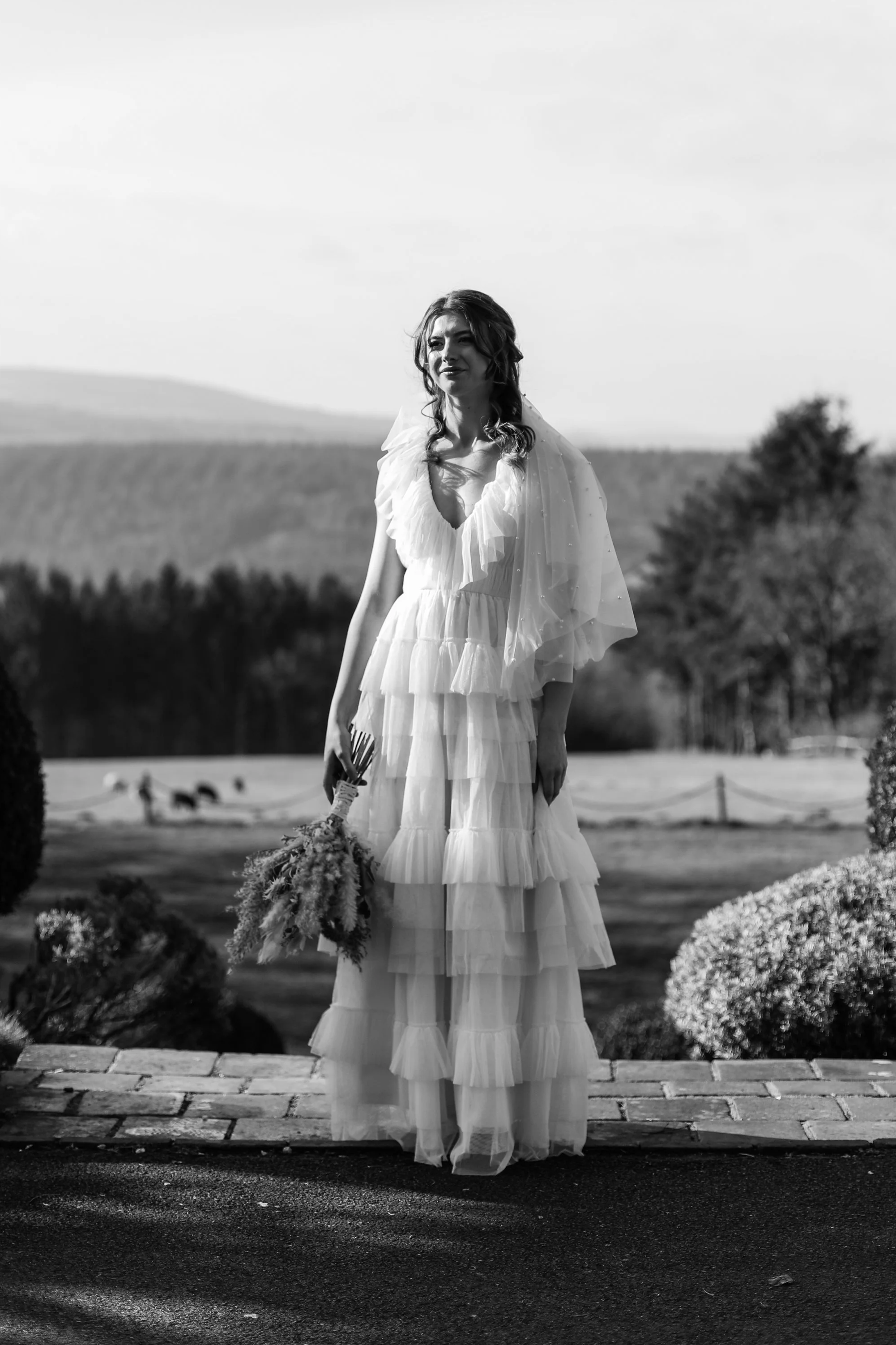 Black and white photo of a woman in a layered, ruffled wedding dress holding a bouquet outdoors with trees and hills in the background.