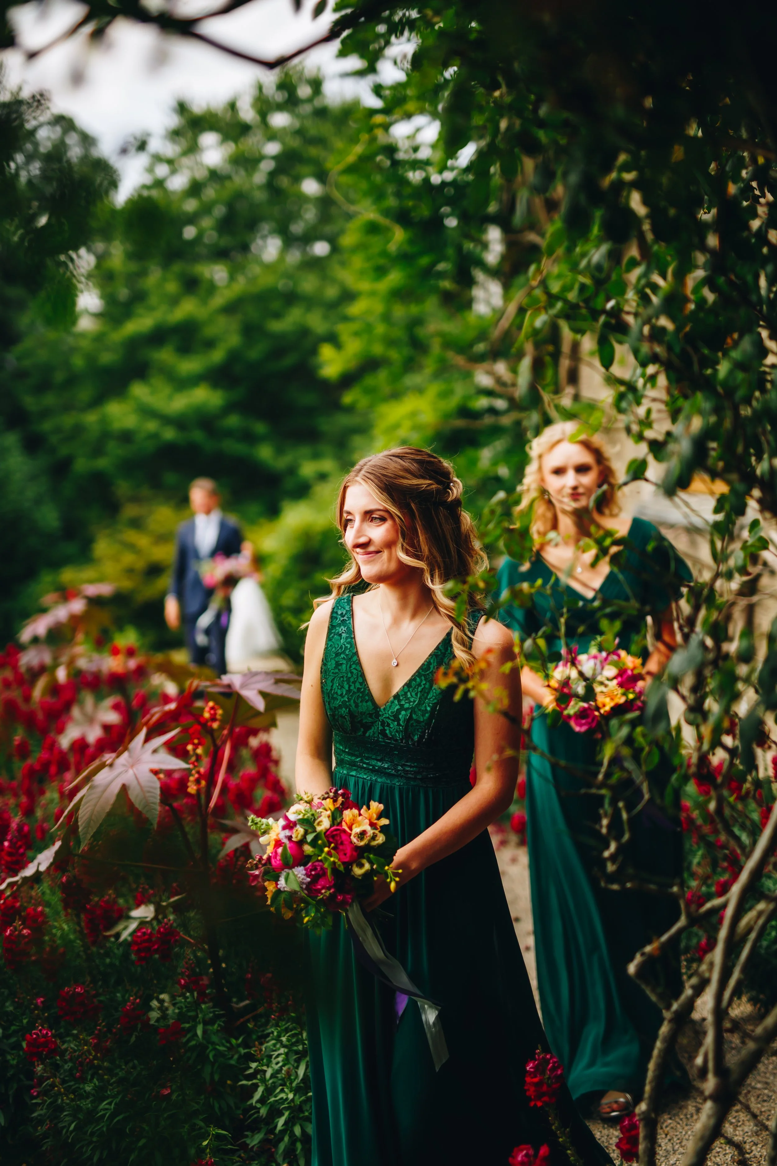 A woman in a green dress holding a bouquet, standing among flowers with a man and another woman in the background outdoors during a wedding ceremony.