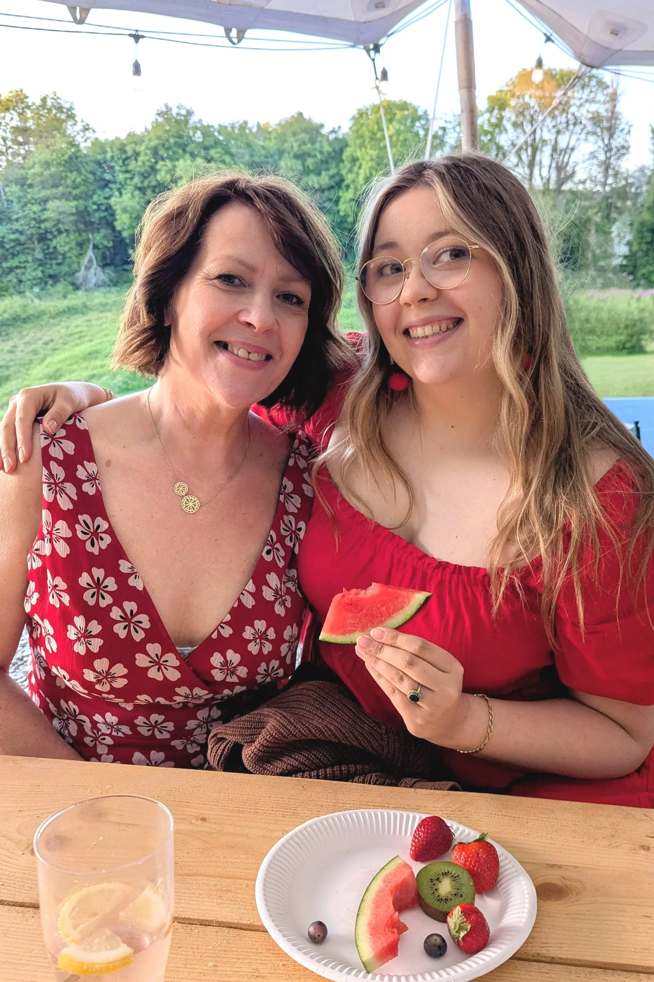 Two women smiling at a camera, one holding a slice of watermelon, sitting at a table with a plate of assorted fruits and a glass of water with lemon slices, outdoors with greenery in the background.