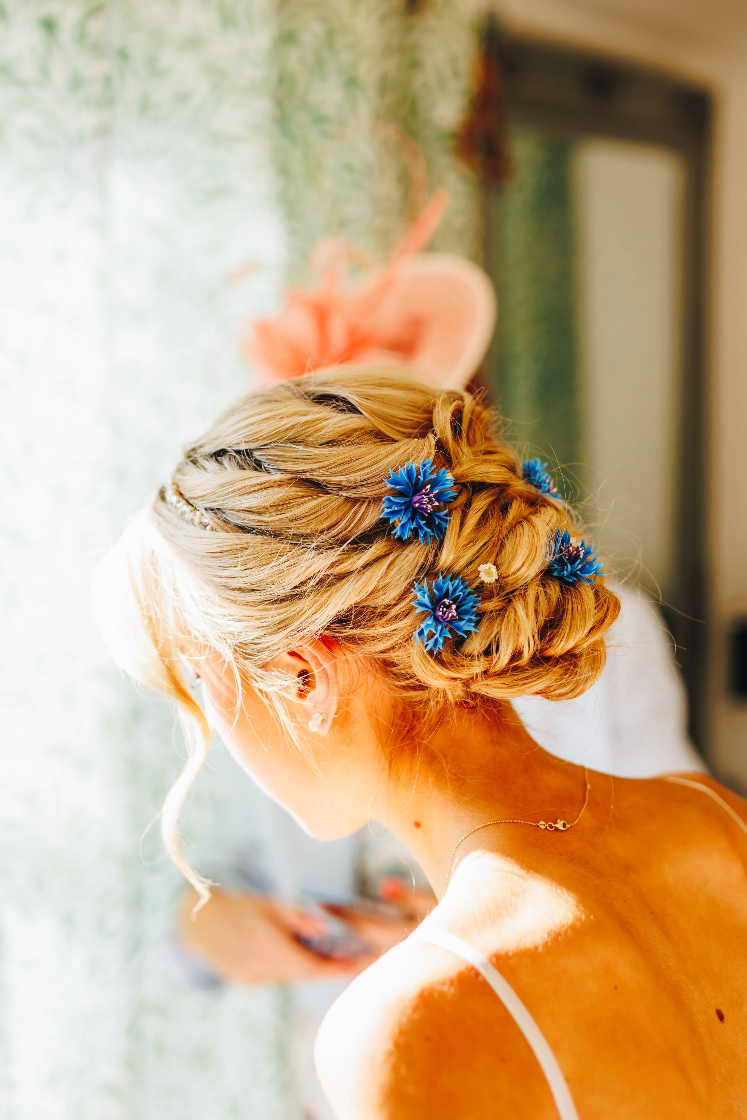 A woman with blonde hair styled in an elegant updo adorned with blue flowers, wearing a delicate necklace and earrings, looking down as she prepares for a special occasion or event.