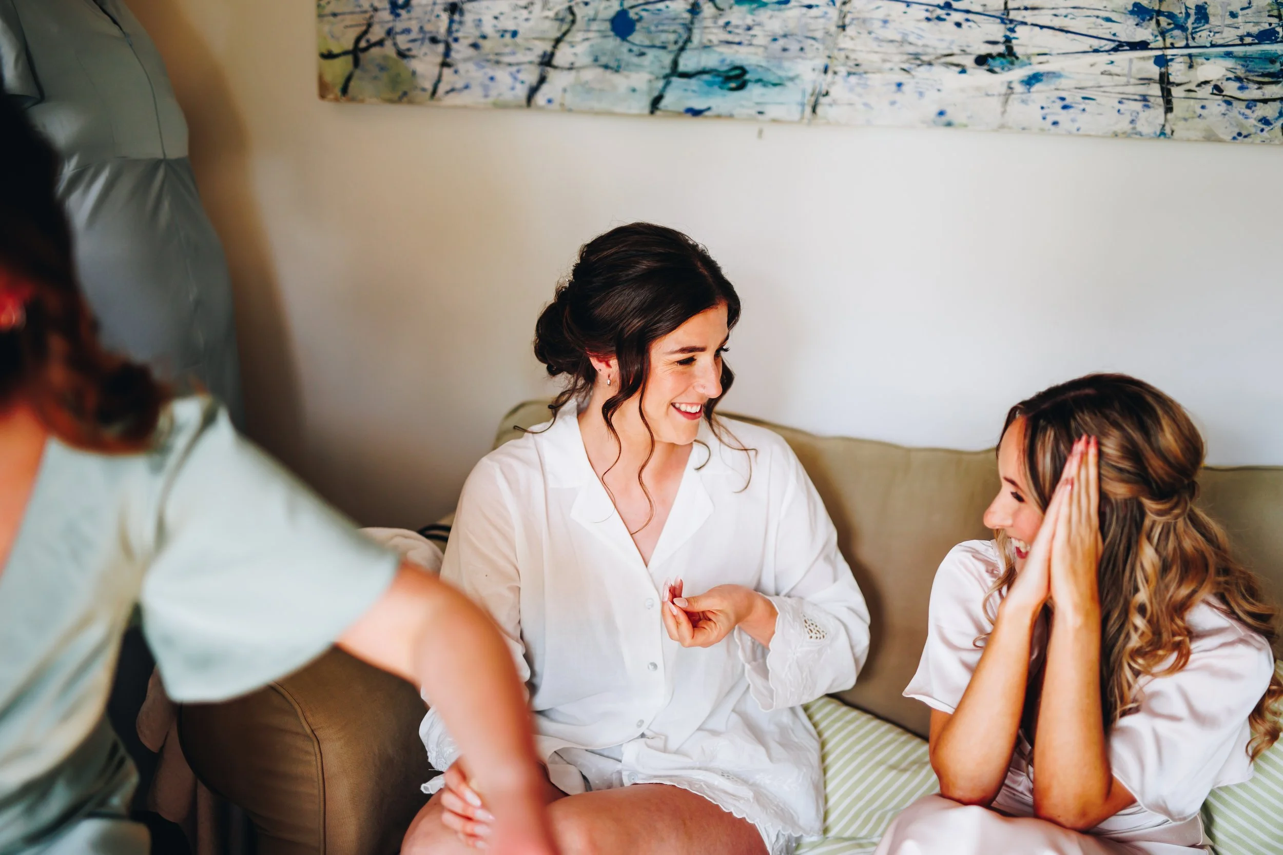 Three women in a room, one woman with dark hair in a white shirt sitting on a couch, two others in pajamas sharing a joyful moment.